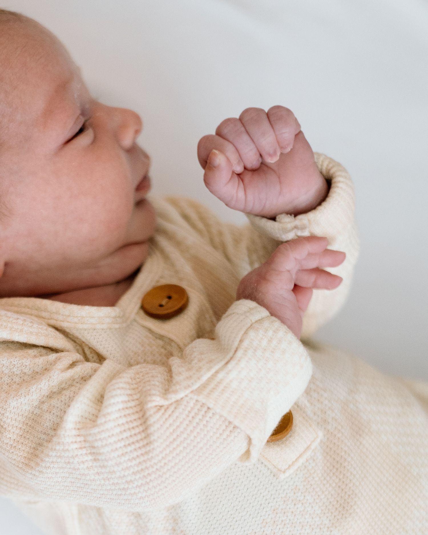 Newborn baby in cream knit cardigan with wooden button, tiny fists raised, peaceful expression.