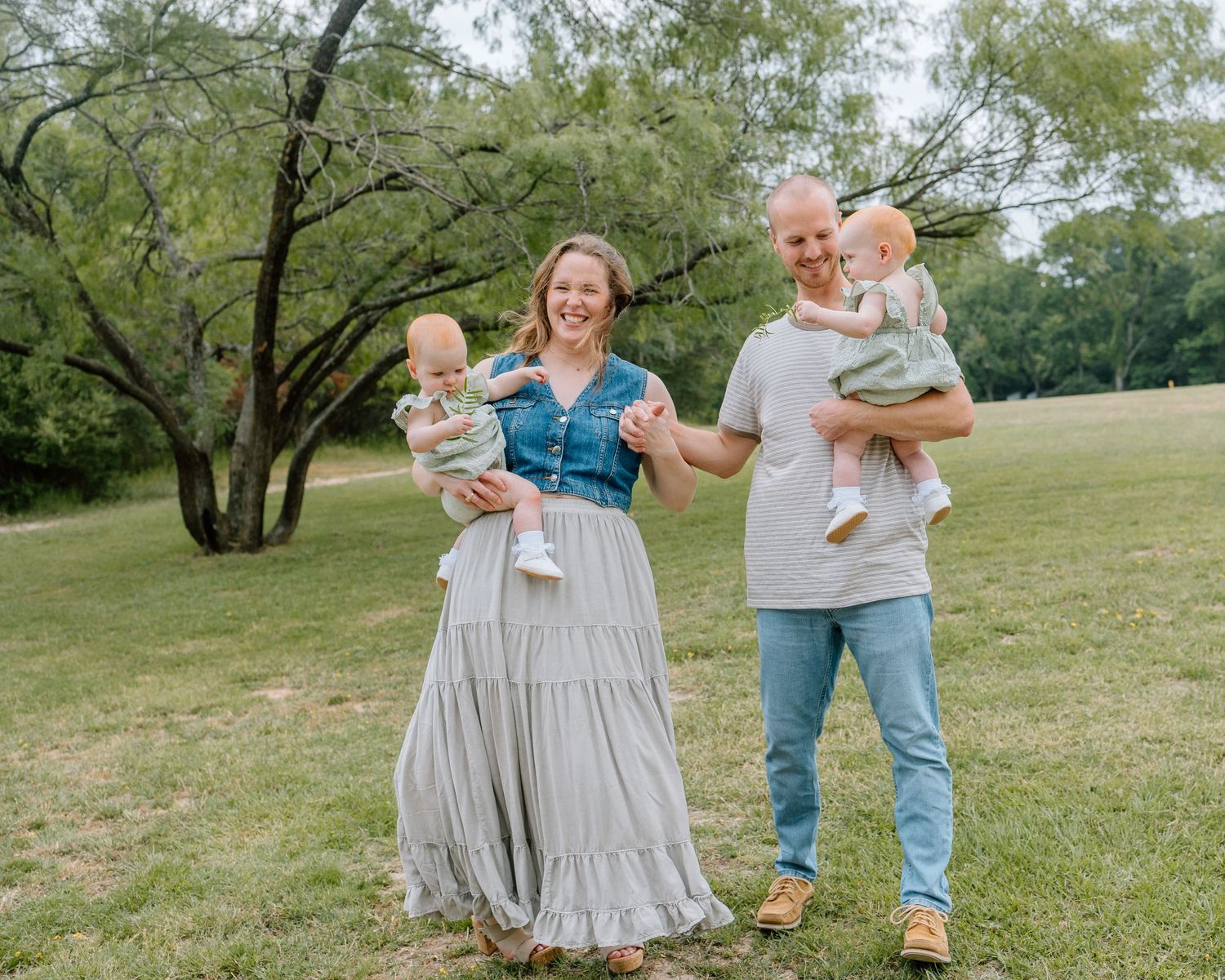 Couple stands together on grassy field near trees on sunny day while each holding small children.