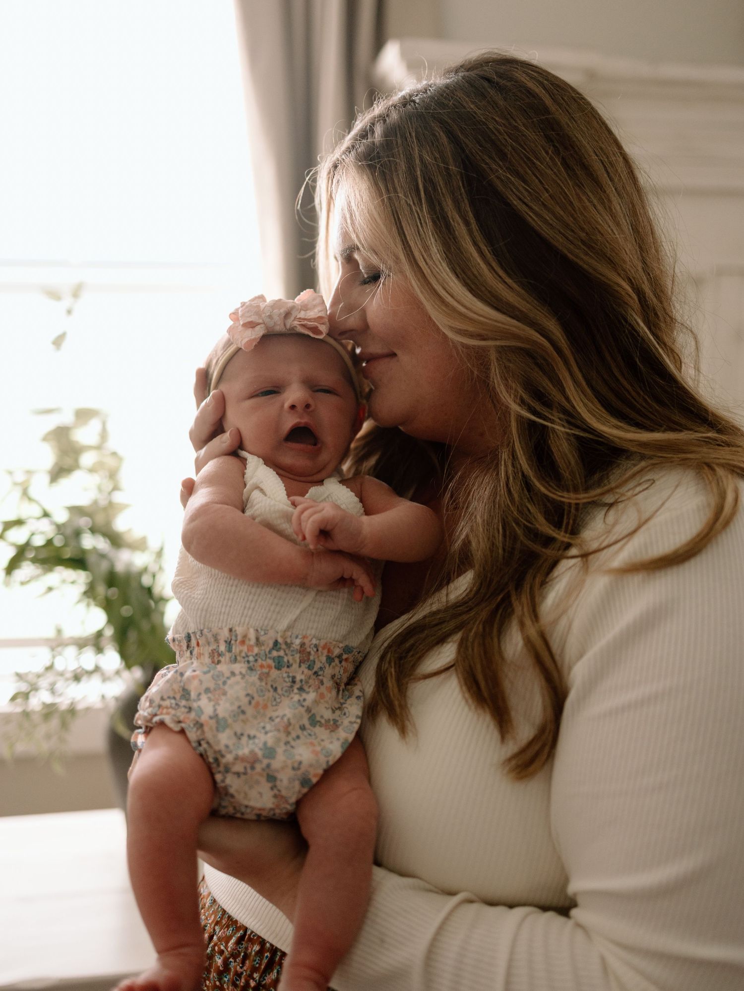 A tender moment as a woman in white cradles and kisses a yawning newborn baby wearing floral clothing by a window.