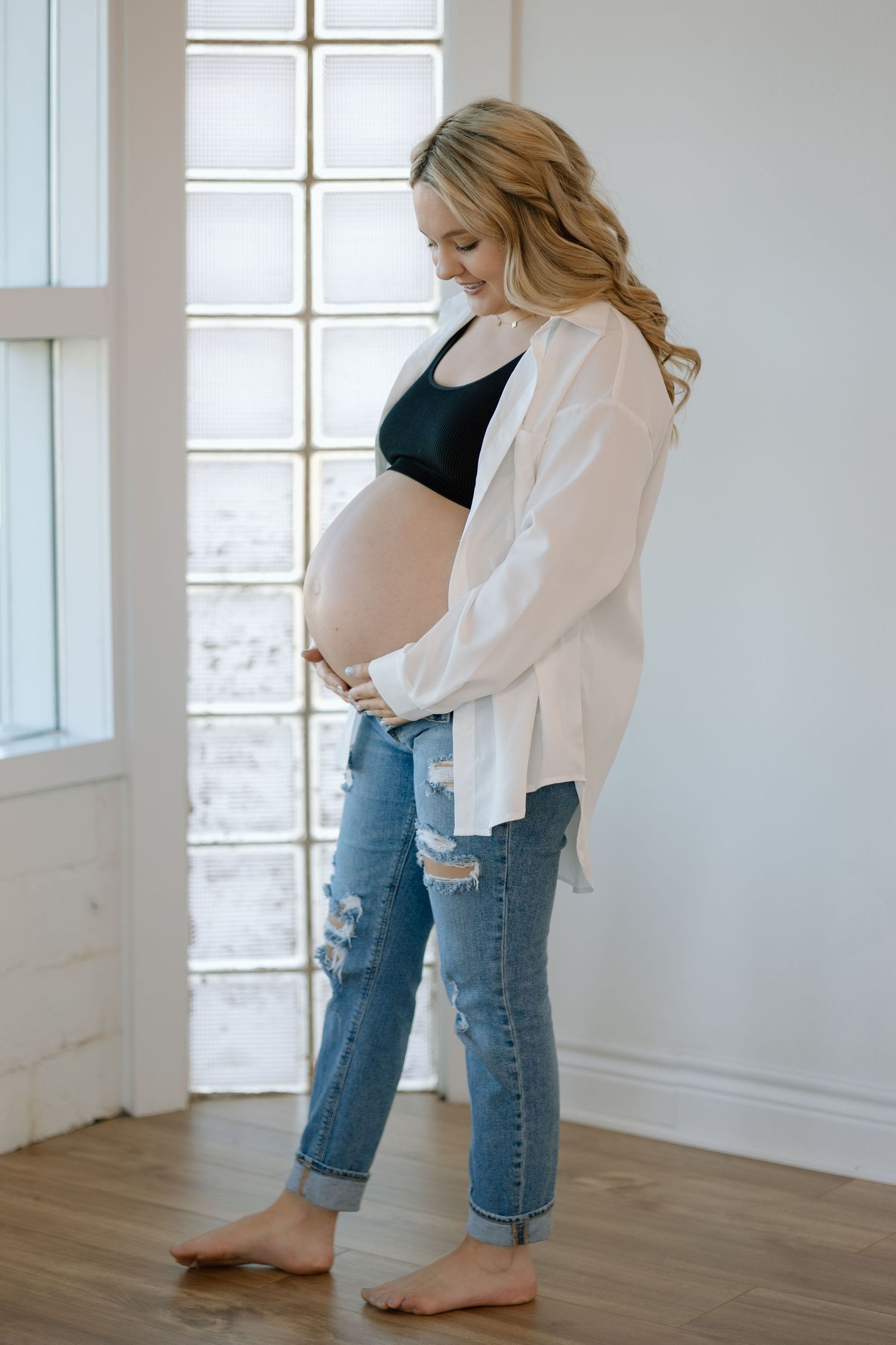 Pregnant woman in ripped jeans and white button down shirt stands barefoot by bright window in minimalist room.
