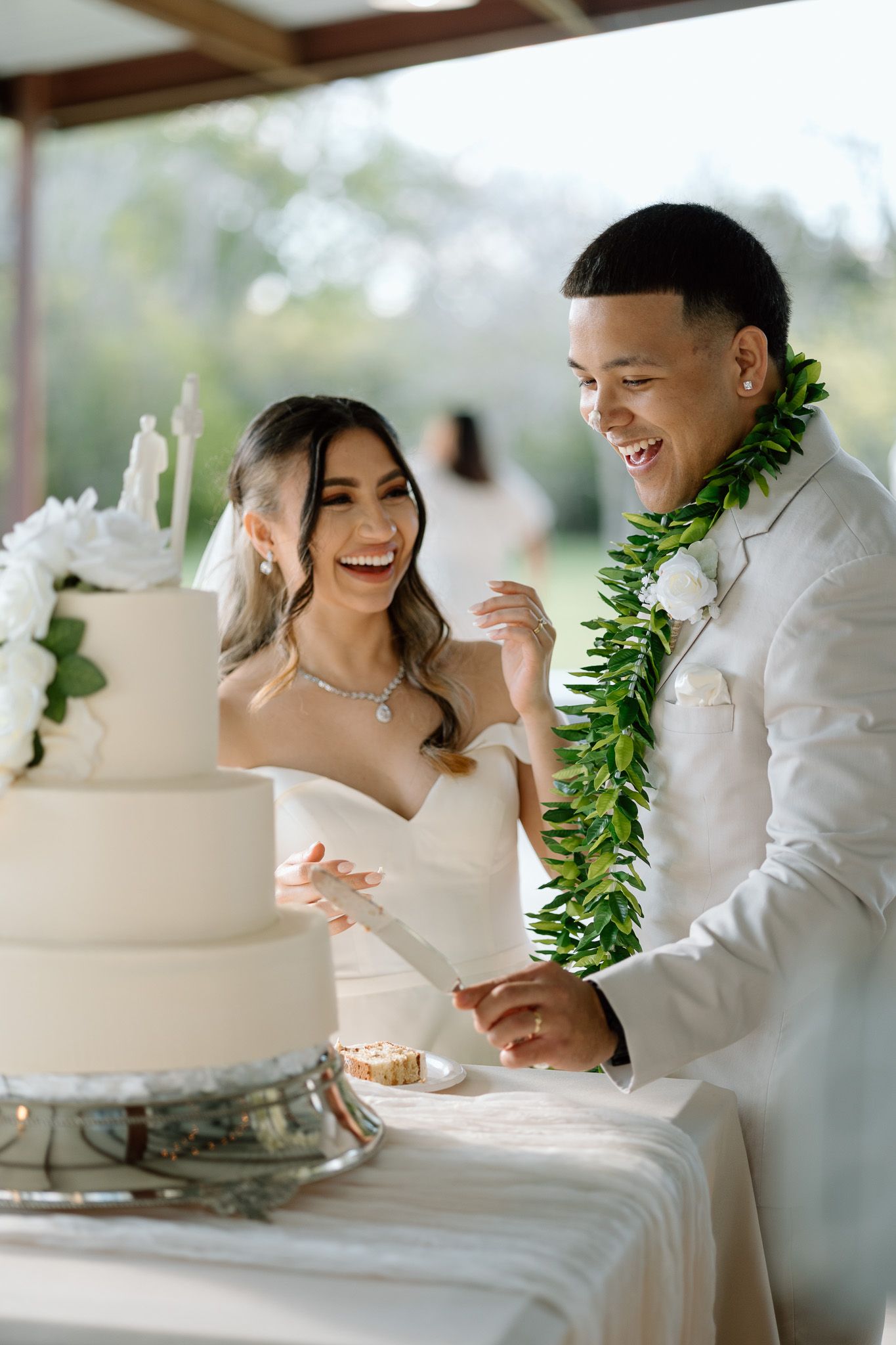 Happy couple laughs while cutting their elegant white wedding cake at outdoor reception.