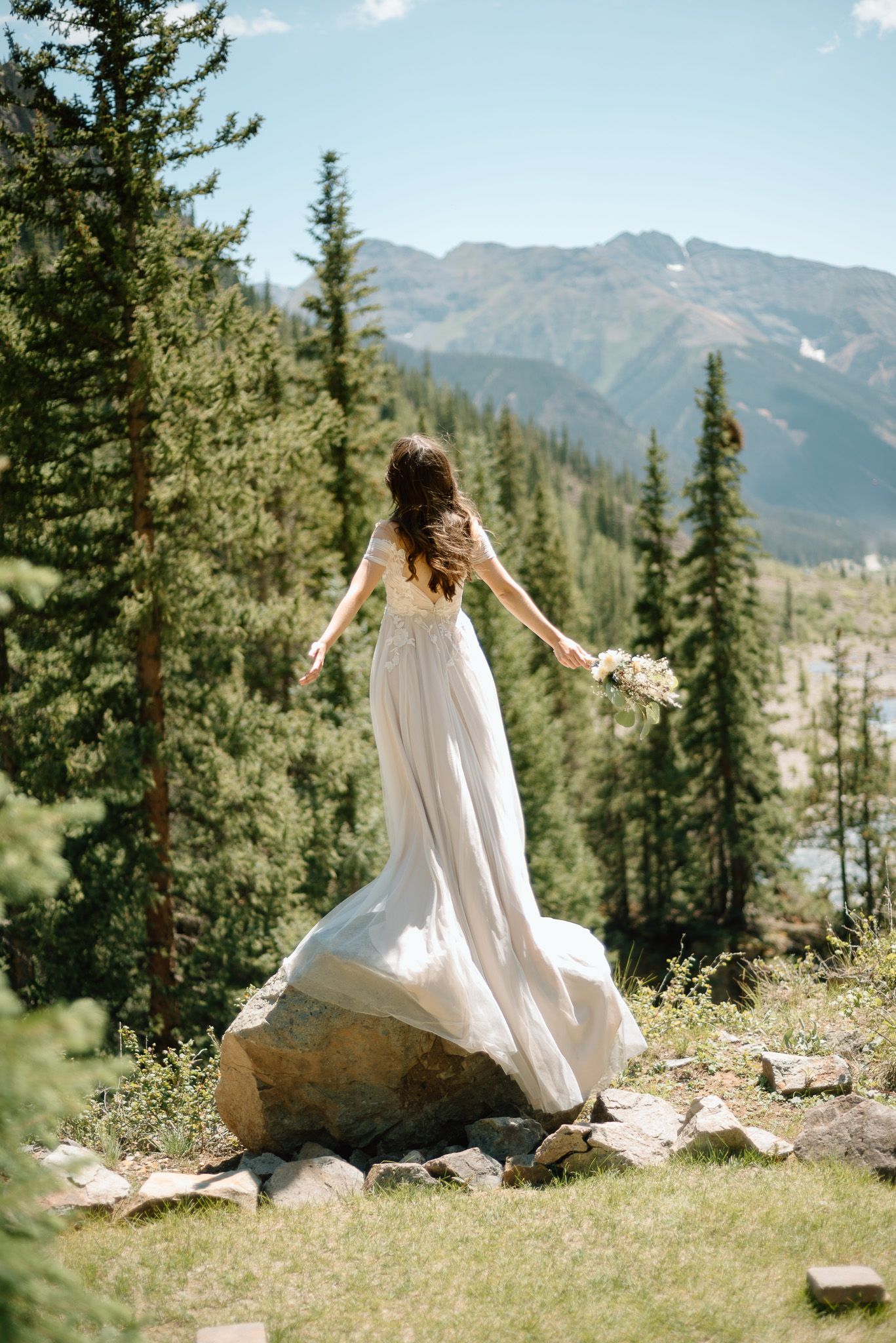 Bride in flowing dress poses with arms outstretched on mountain summit.