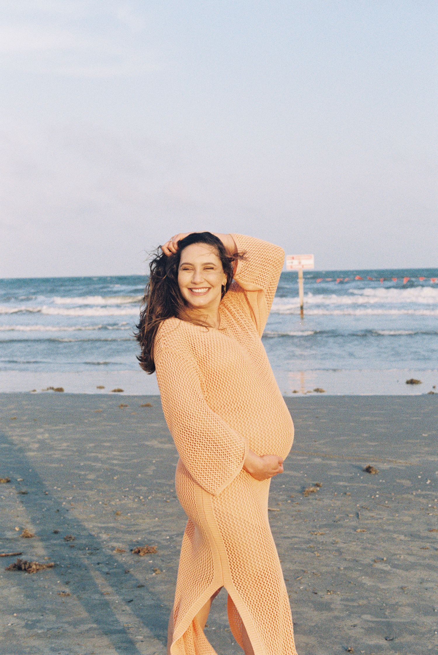 An expectant mother in a peach sweater dress poses joyfully on the beach.