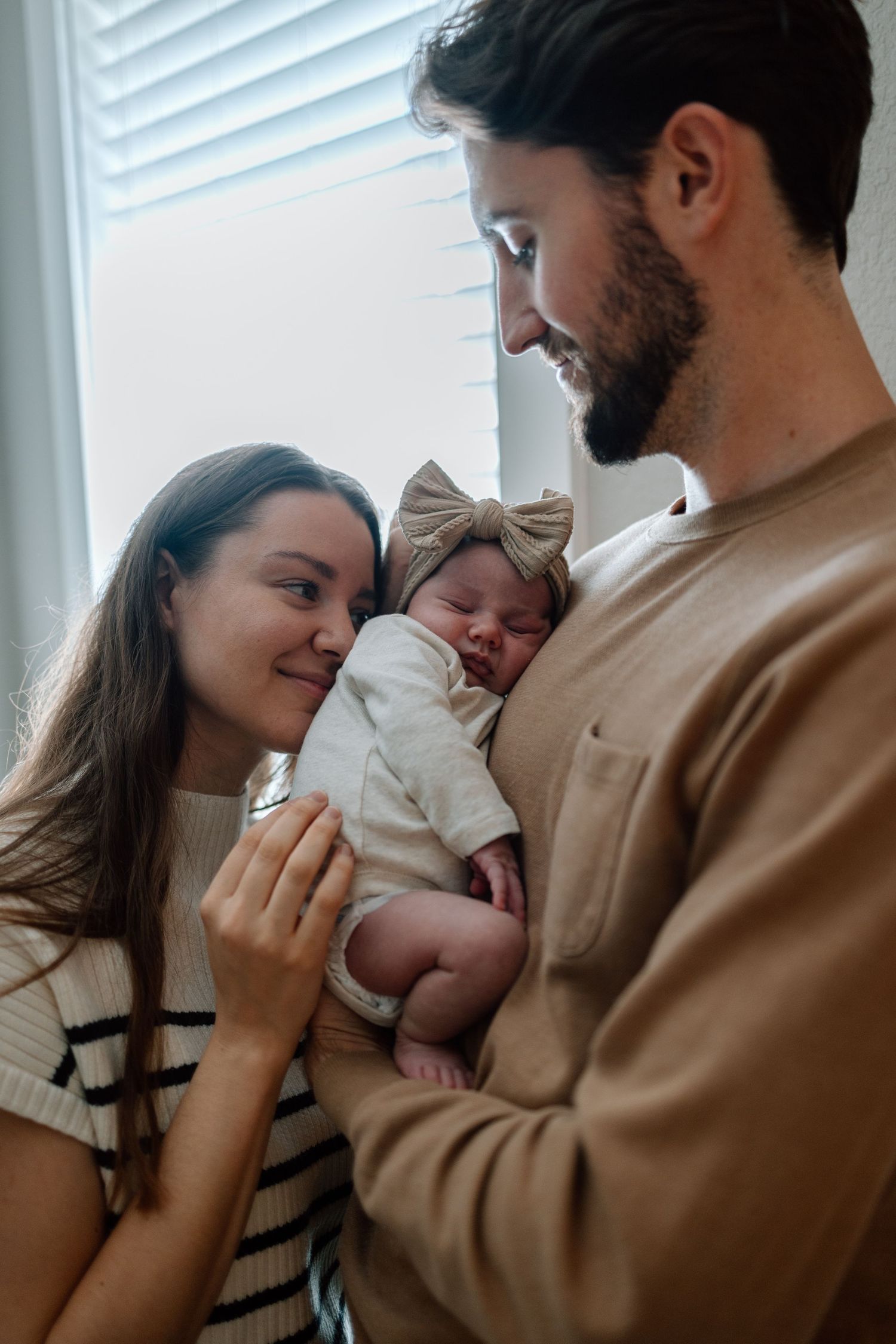 New parents sharing an intimate moment while holding their sleeping newborn baby wearing a bow headband.