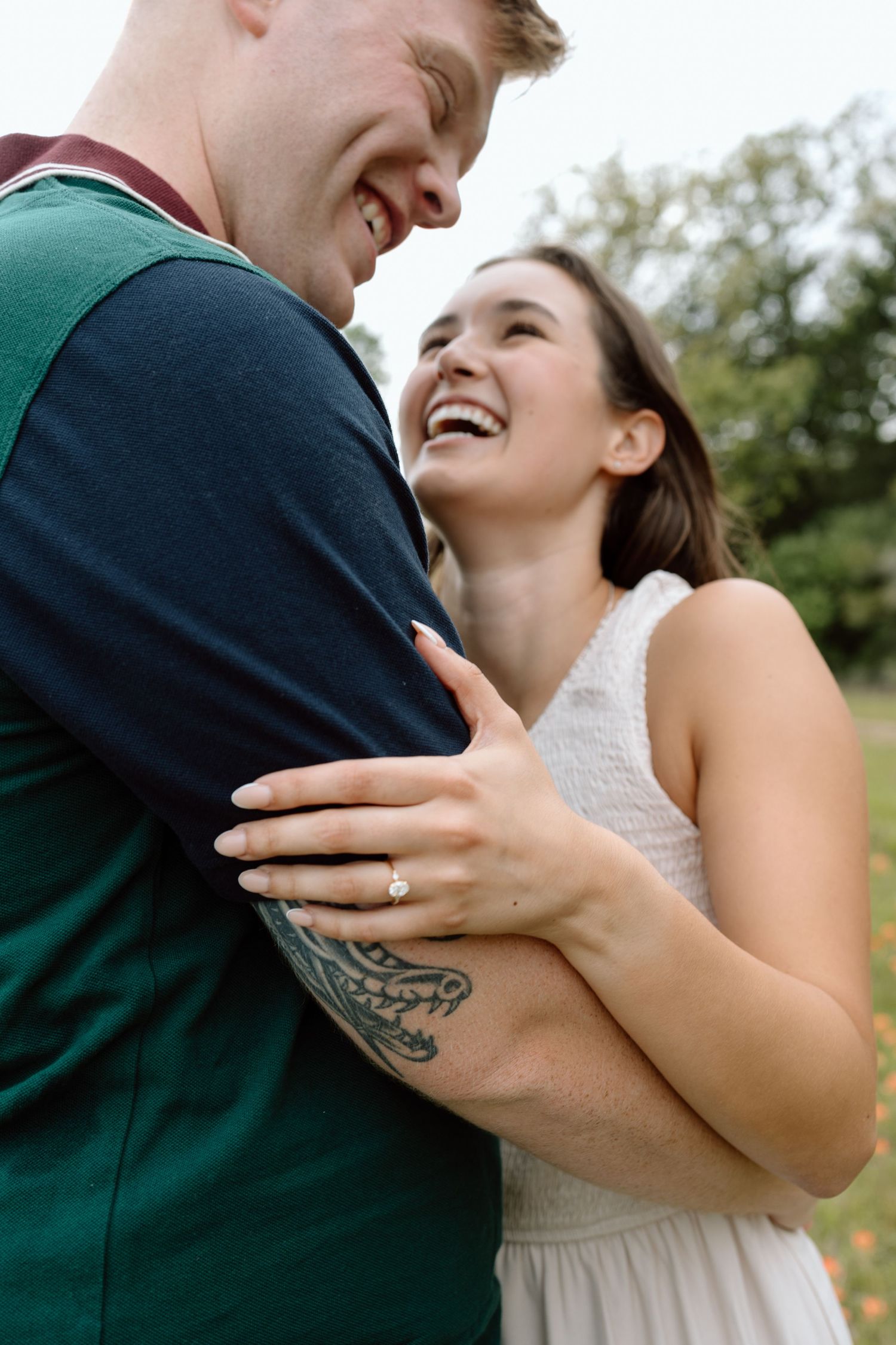 A couple shares a joyful moment as they embrace outdoors, with a tattoo visible on his arm and her wearing a white dress.
