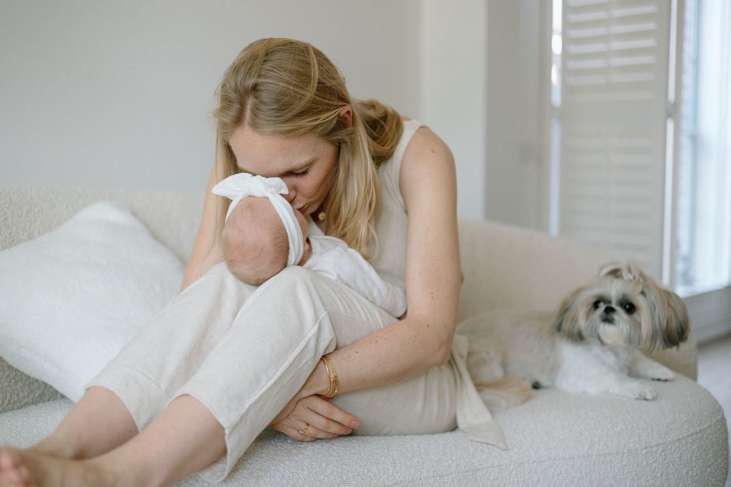 Mother cuddling with infant on white couch while small white dog rests nearby.