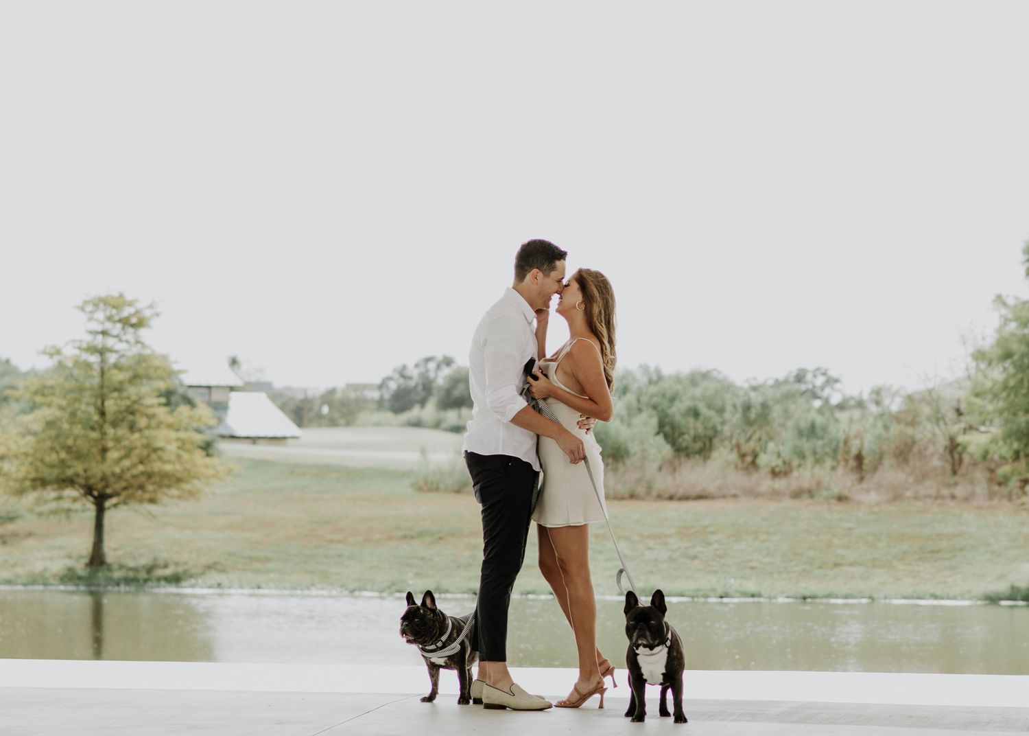 A couple embraces while standing with their two dogs on a concrete path near a lake.