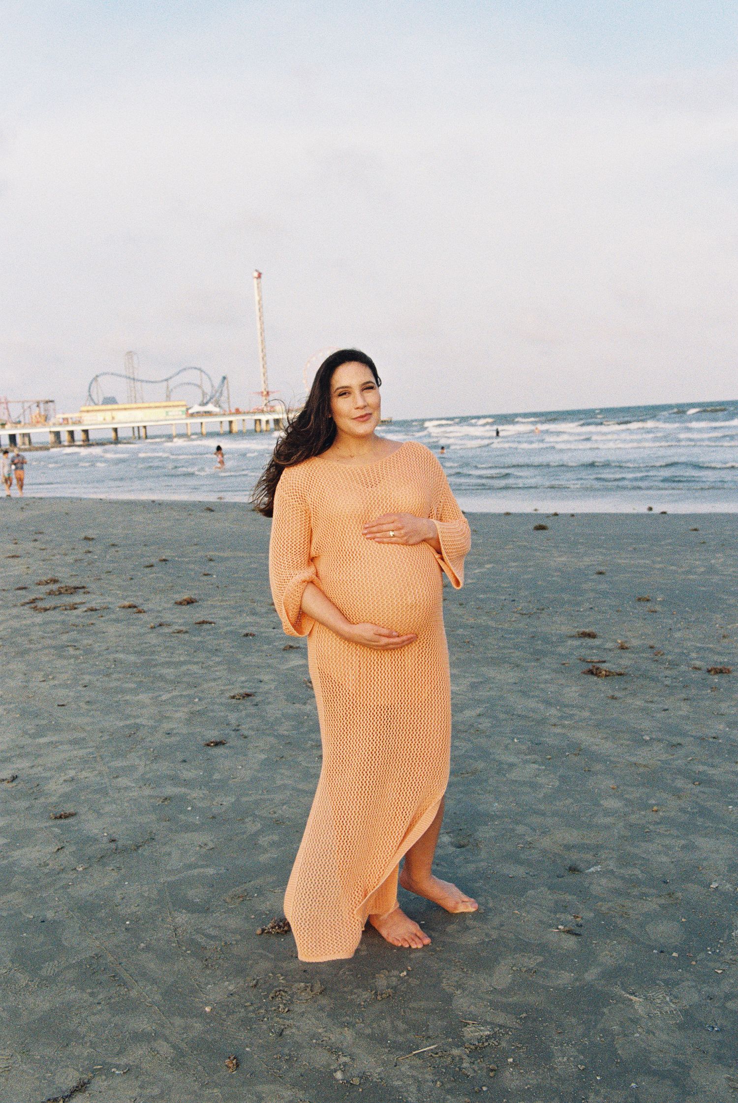 Maternity portrait of woman in flowing peach dress standing on beach with pier in background.