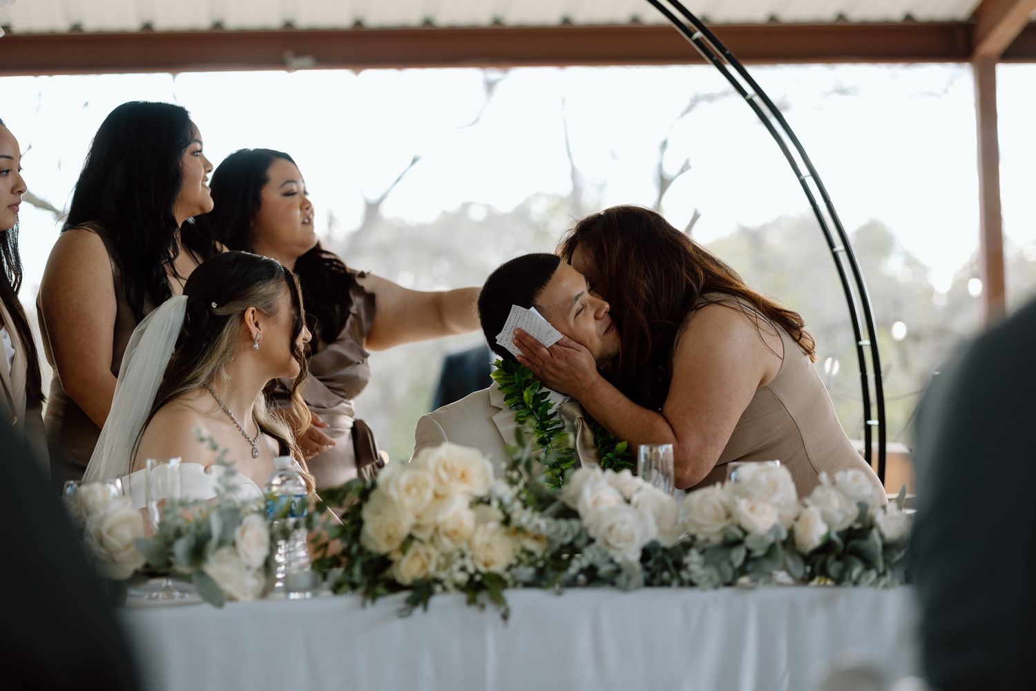 Wedding reception scene with guests gathered at a decorated table under covered outdoor space.
