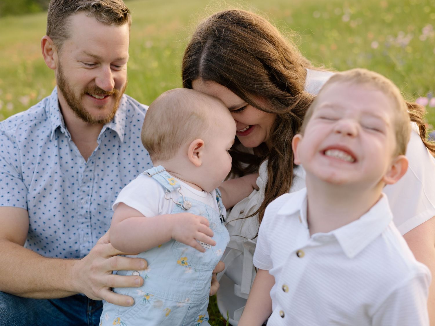 Joyful family sits together outdoors in grass while baby looks on and child laughs during candid moment.