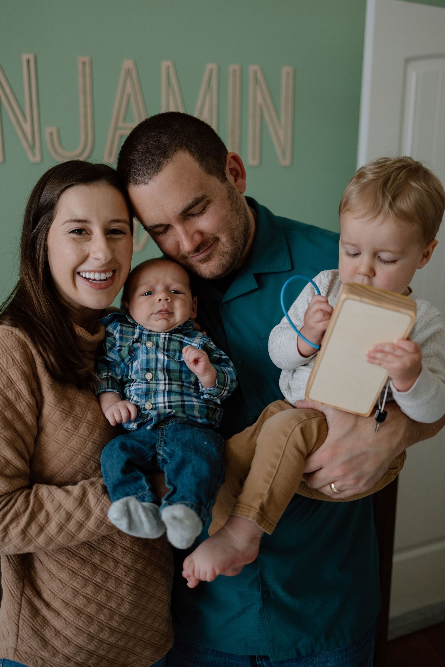 A happy family moment captured indoors as parents hold their two young children against a light colored wall background.