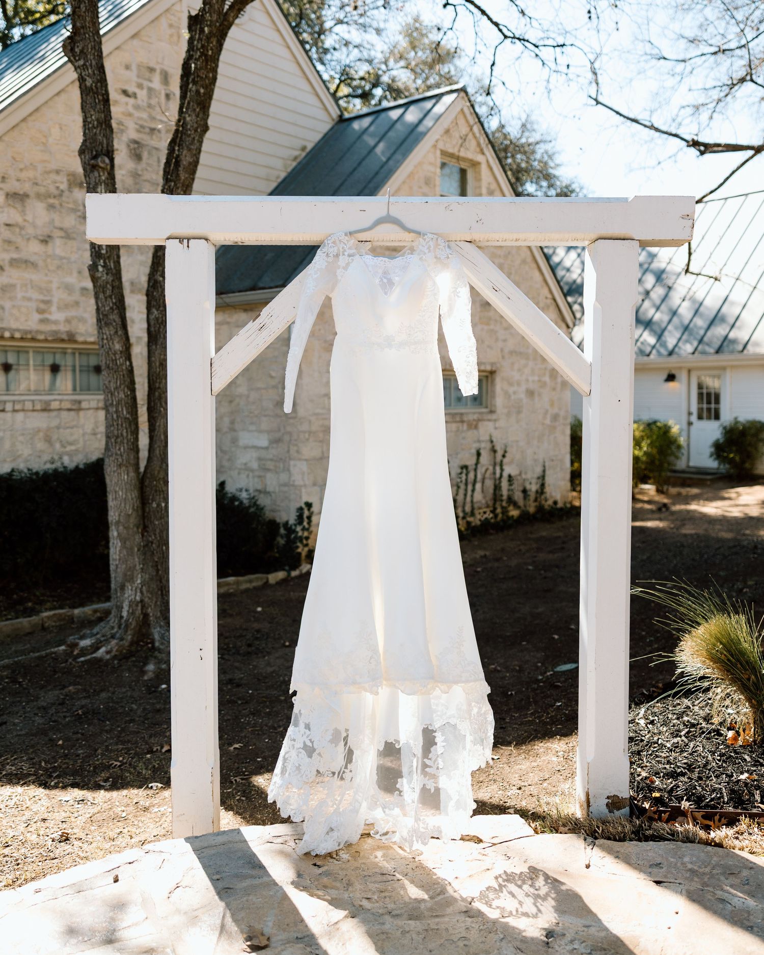 White lace wedding dress with long sleeves hanging on outdoor white arch at rustic stone venue.