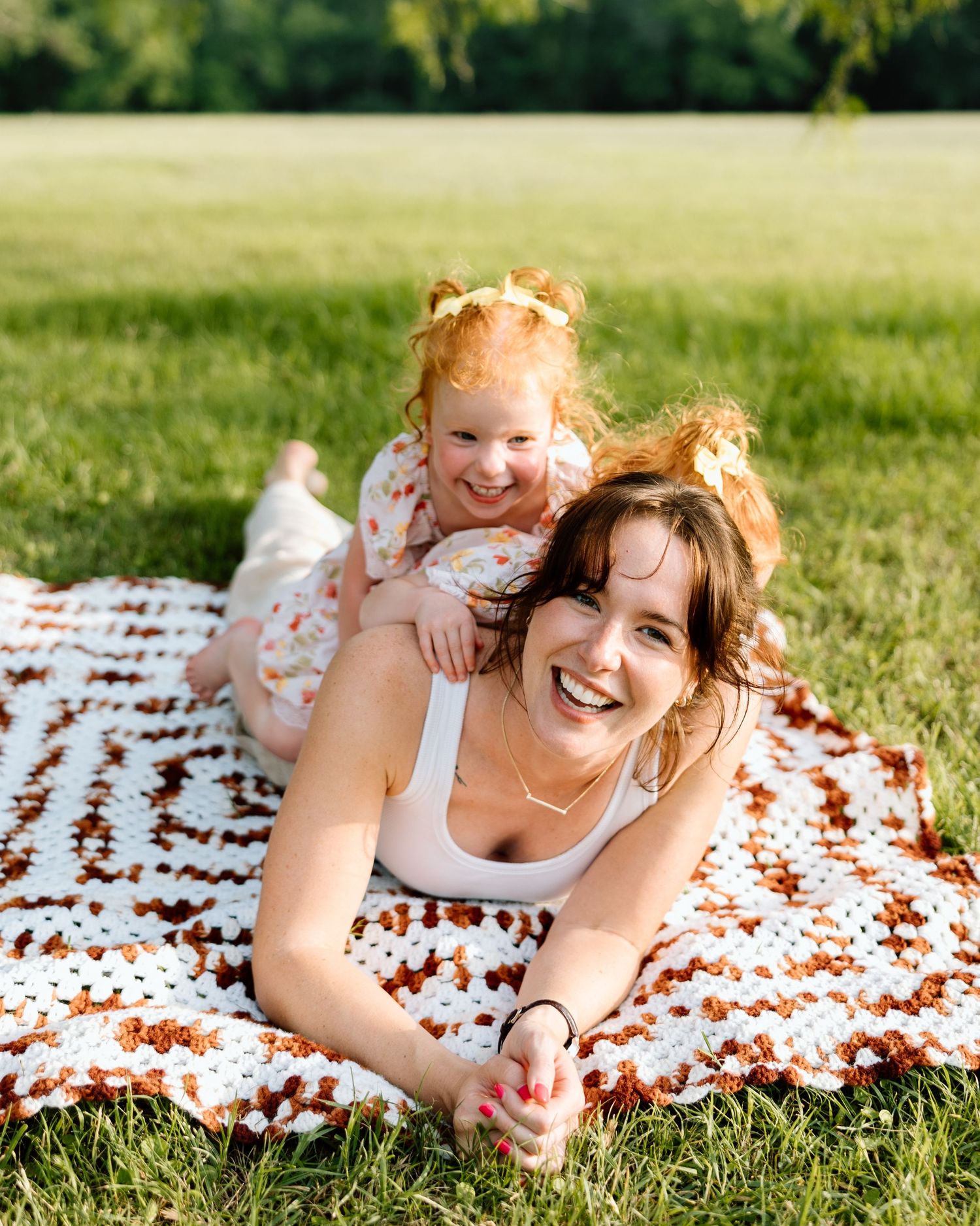 A laughing mother and red-haired toddler girl lying on a crochet blanket in a sunny green park.