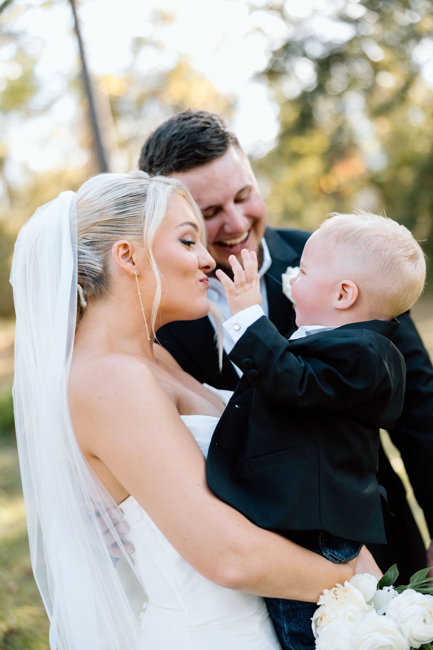 Bride and groom smile joyfully as adorable baby in tuxedo reaches out to touch bride's face.