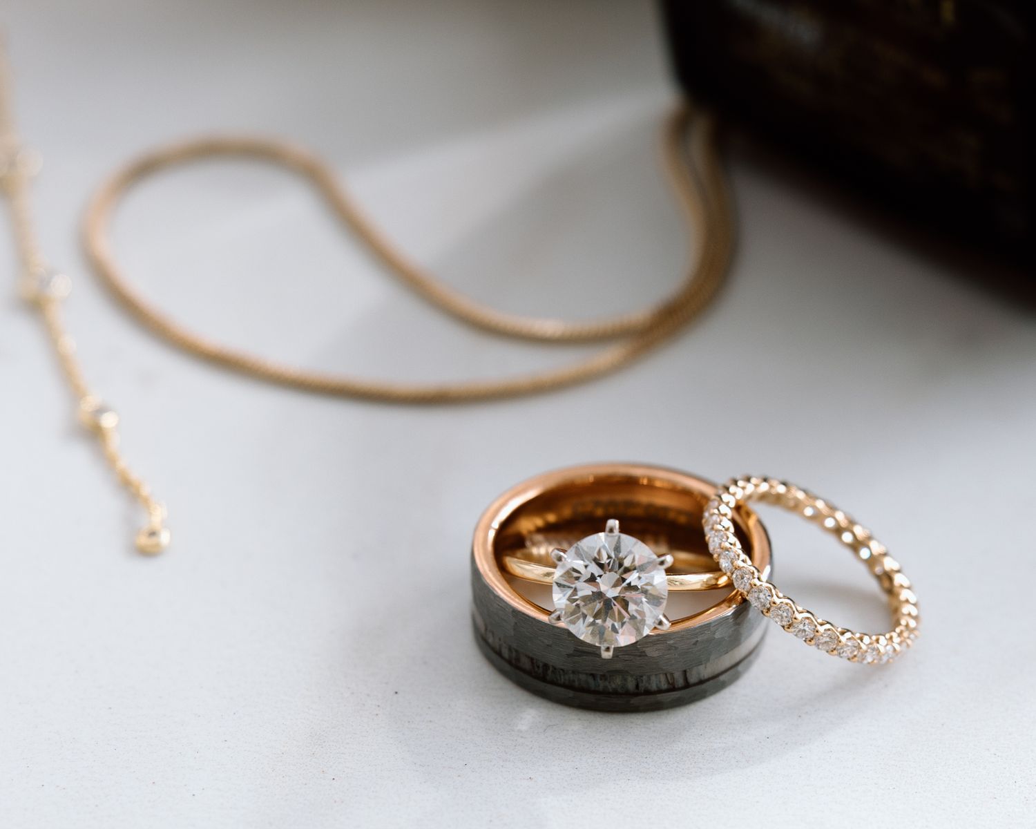 Rose gold diamond engagement ring stacked with wedding bands and necklace on white surface.