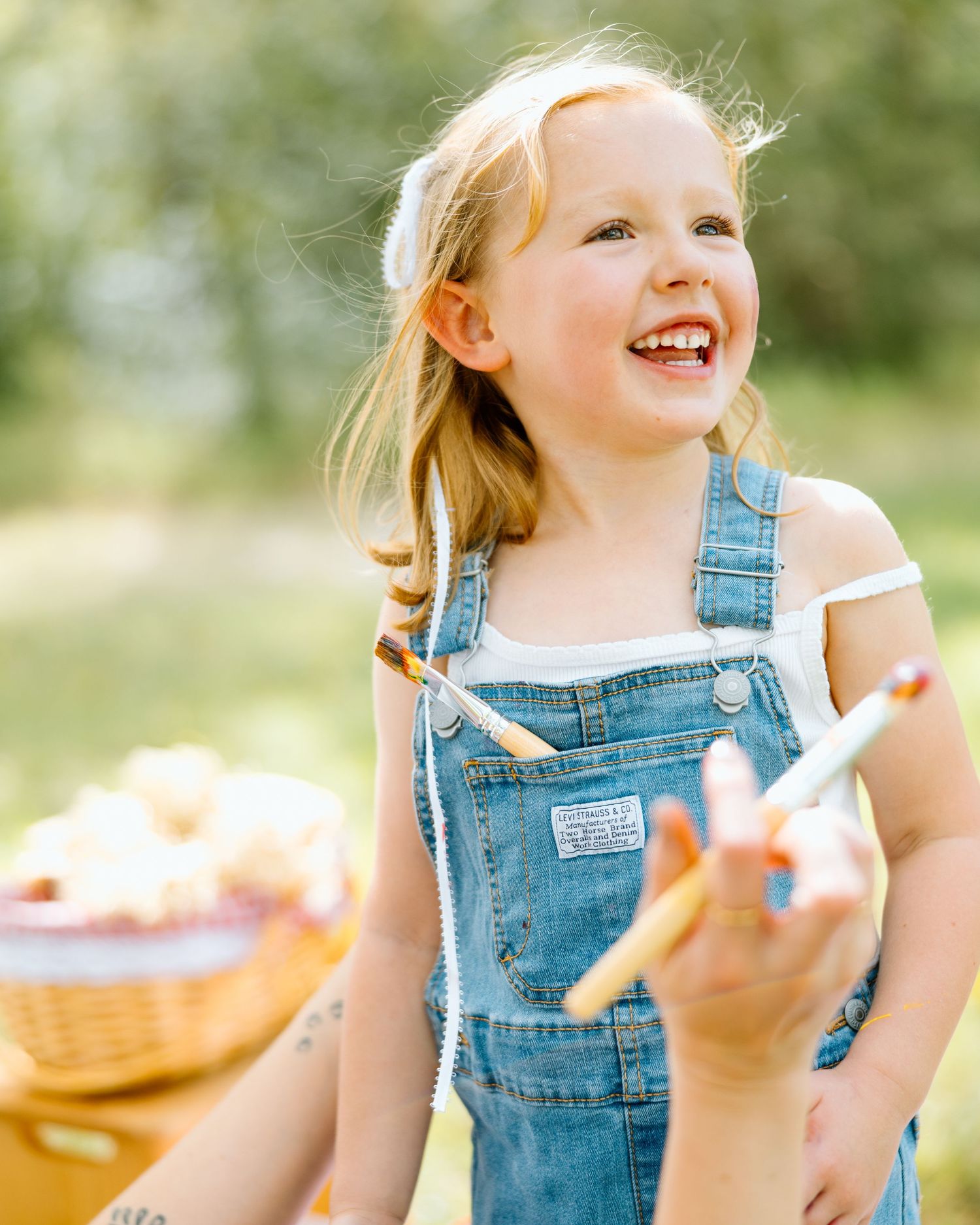 A joyful young girl in denim overalls laughs outdoors, holding a breadstick near a wicker basket.
