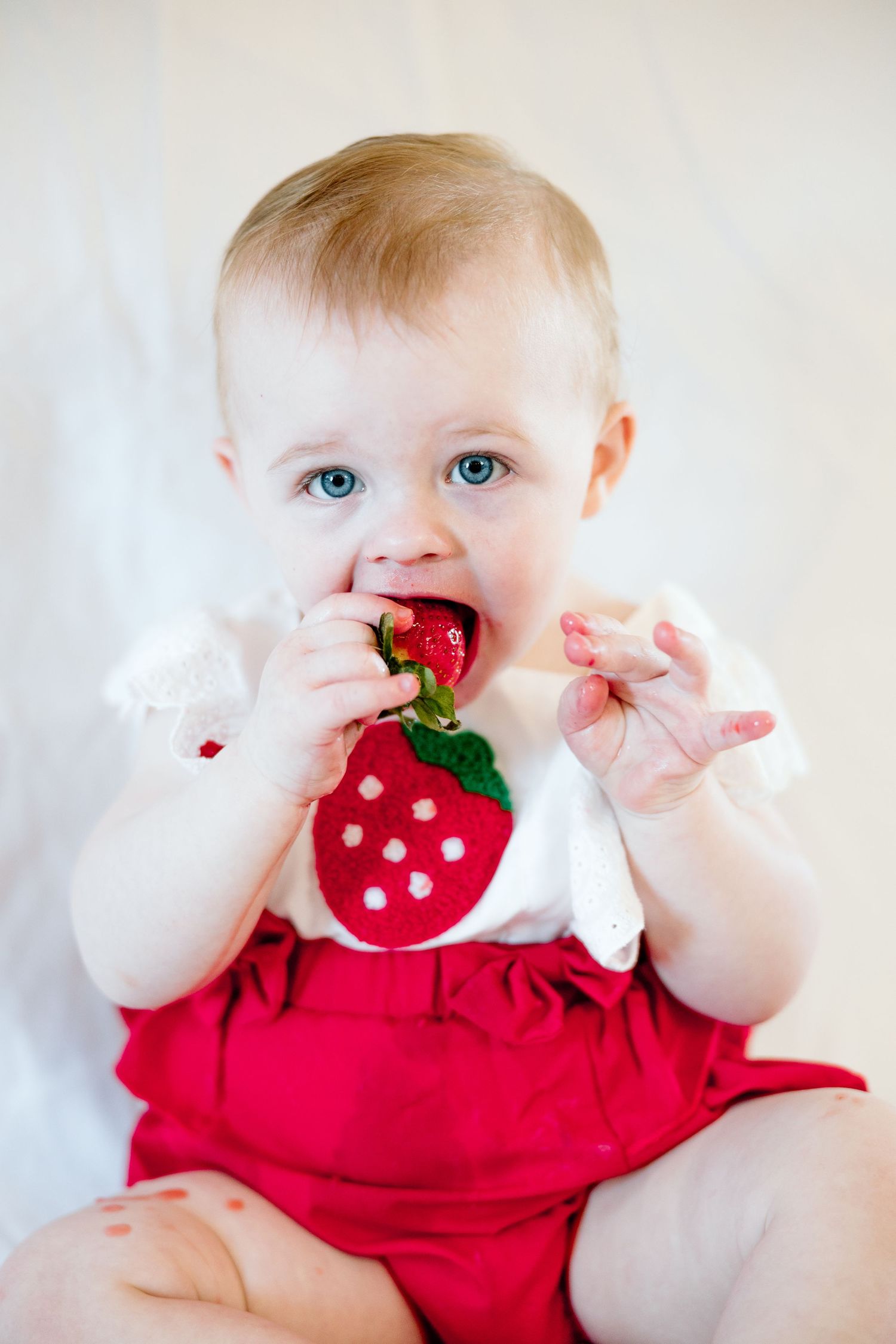 Adorable blue-eyed baby in red strawberry romper biting into a ripe strawberry.