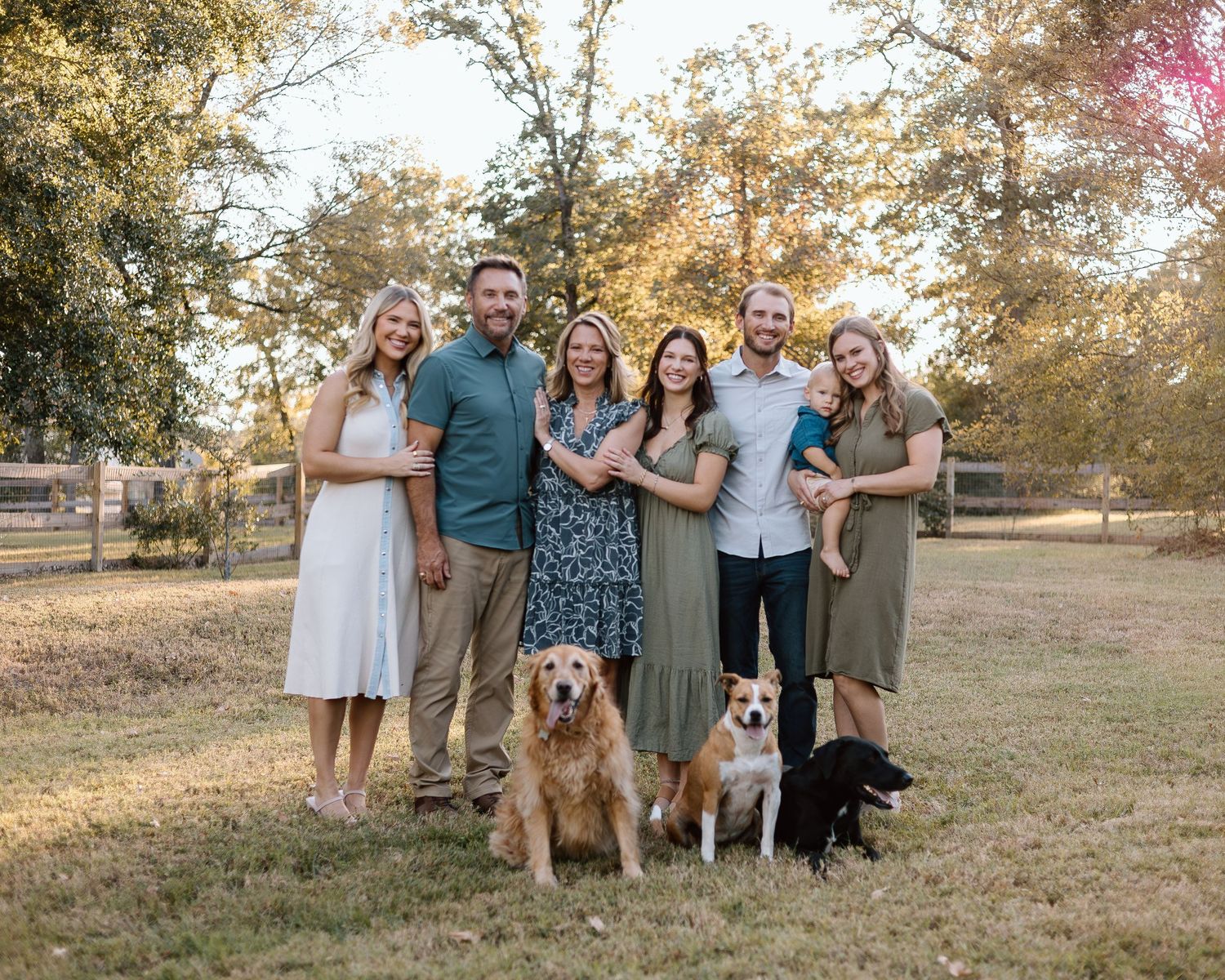 Family of six smiling outdoors with three dogs on a sunny day in a grassy park setting.