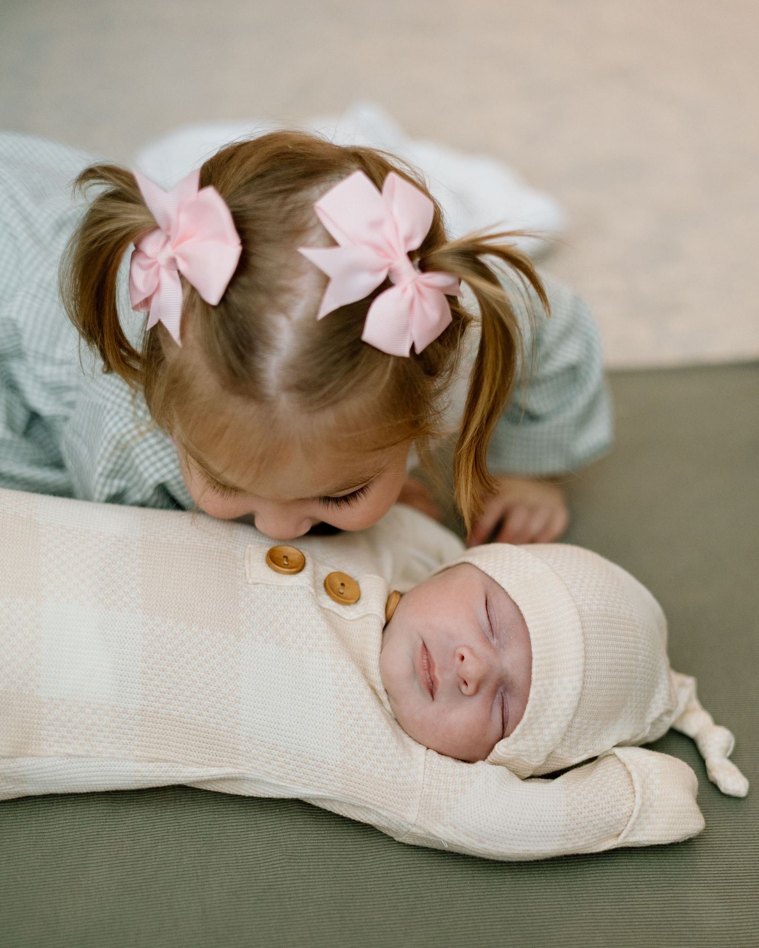 Adorable toddler girl with pink hair bows gently kissing a sleeping swaddled newborn baby sibling.