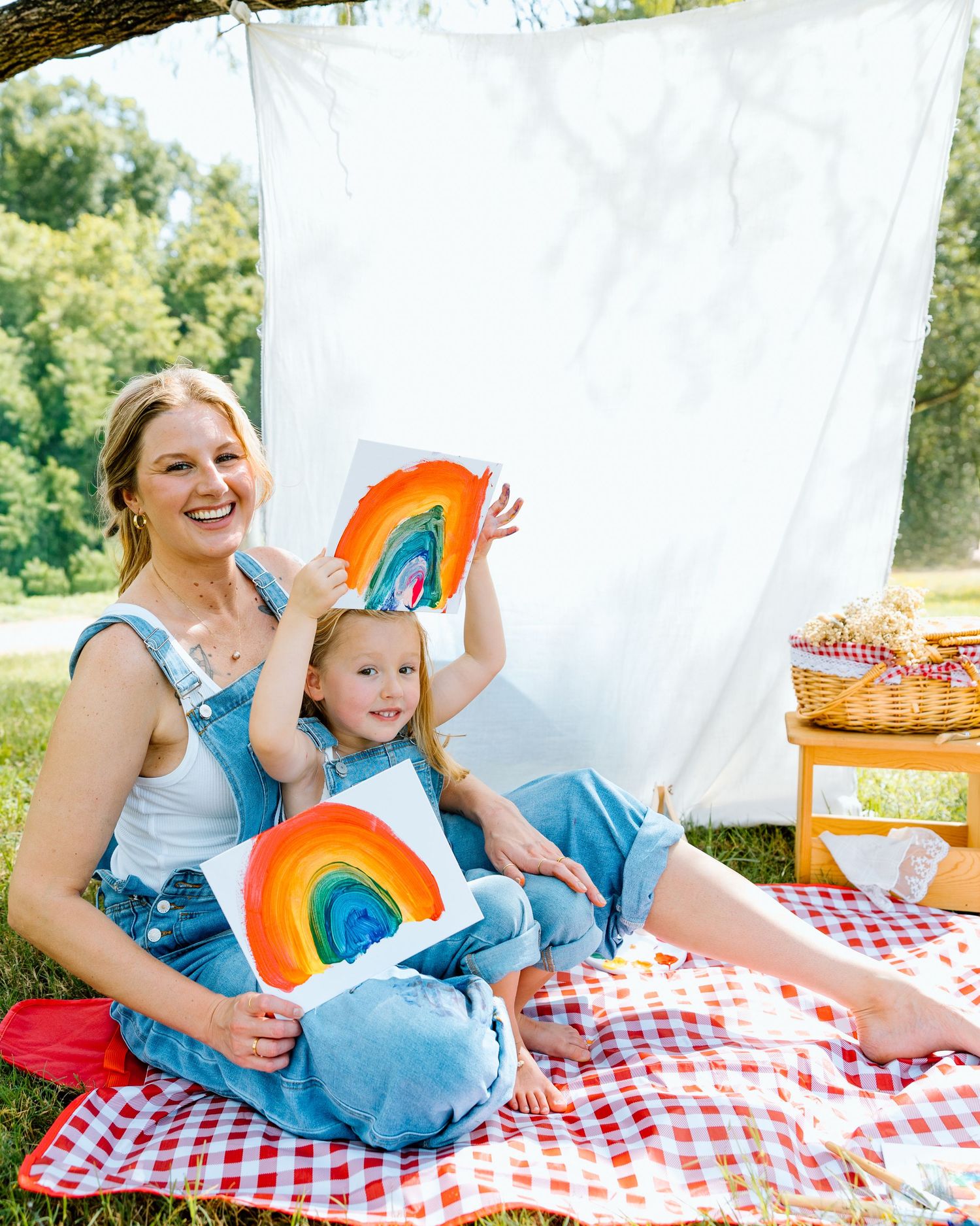 Mom and toddler daughter in matching denim overalls showing rainbow paintings on red gingham picnic blanket outside.