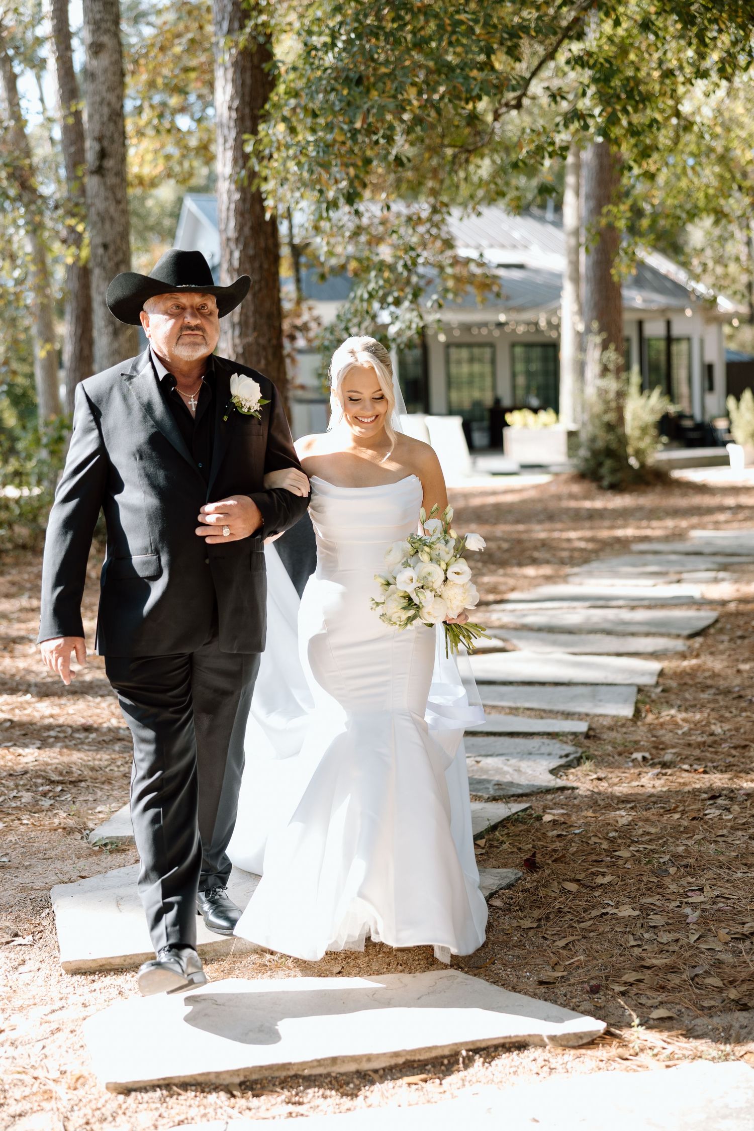 Bride in strapless mermaid gown walks down outdoor path with older man in black suit and cowboy hat.