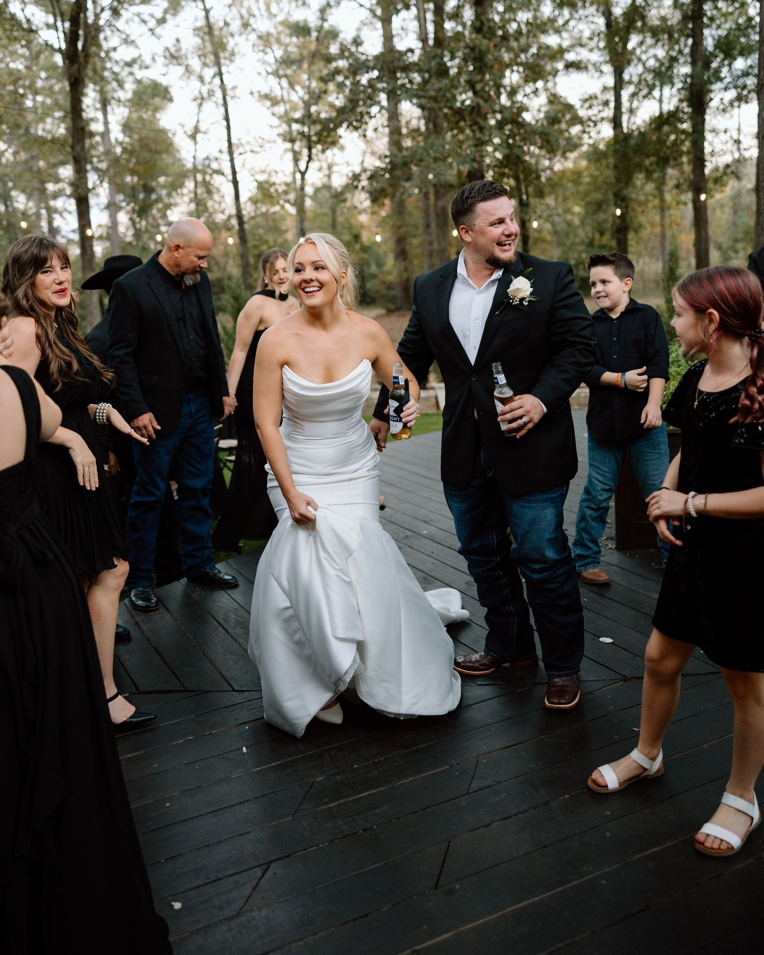 Laughing bride and groom walk through guests on outdoor wooden dance floor at evening wedding reception.