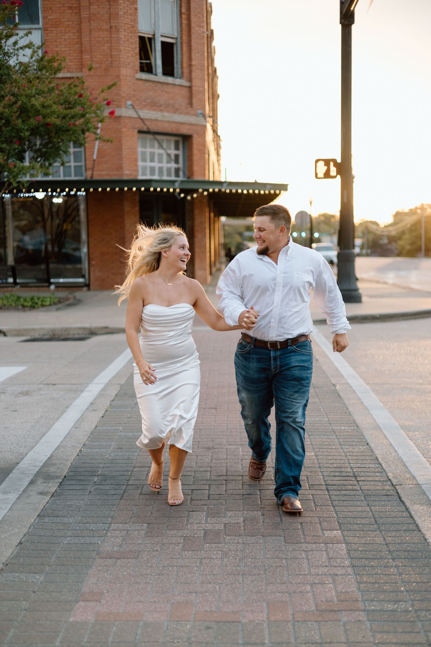 Couple holding hands and laughing while running together on a brick sidewalk at sunset in a charming downtown area.