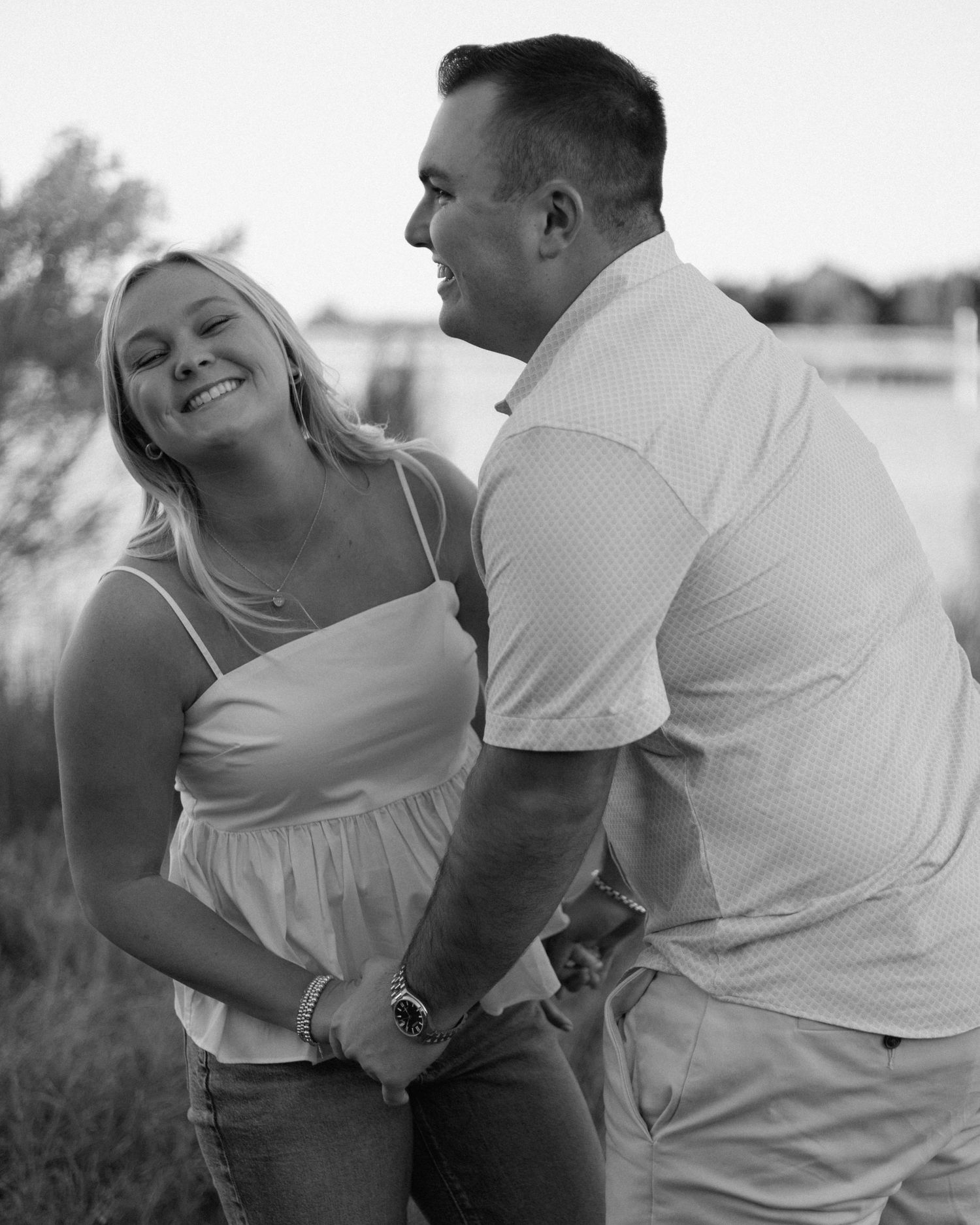 Black and white photo of a smiling couple holding hands outdoors near water, sharing a joyful moment together.
