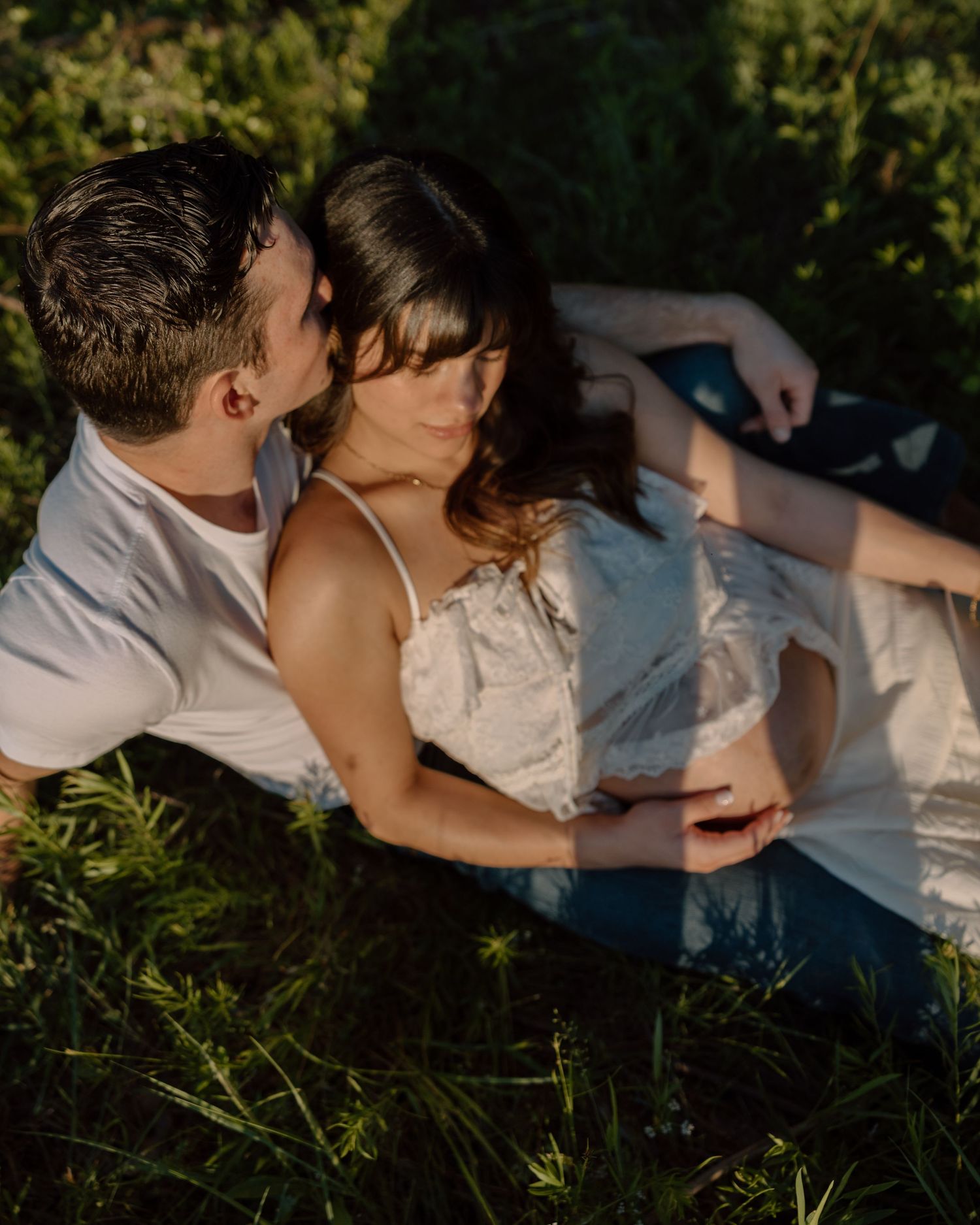Couple laying in green grass sharing a romantic moment outdoors in warm golden sunlight.