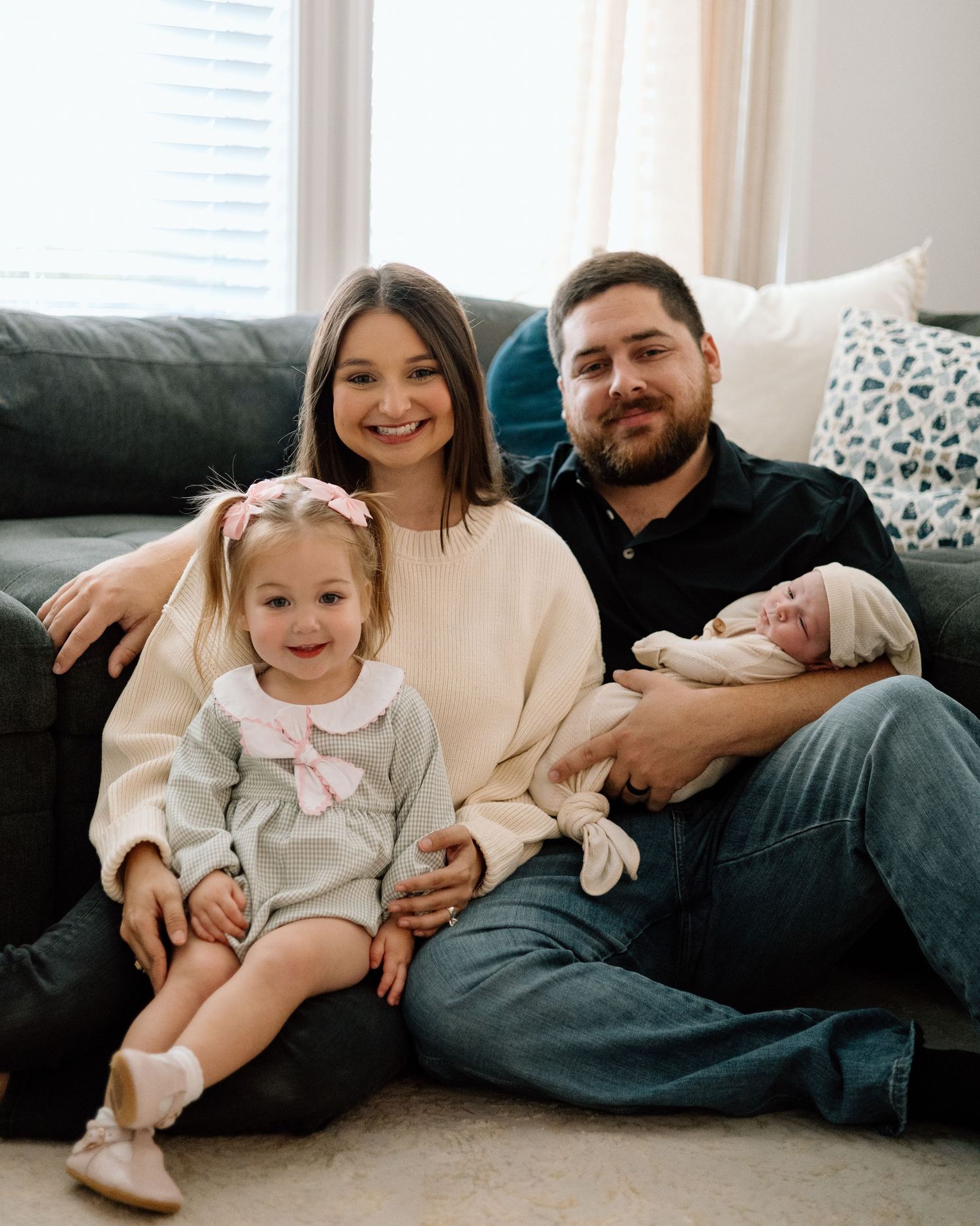 Happy family of four posing together on living room floor, parents with toddler girl and newborn baby.