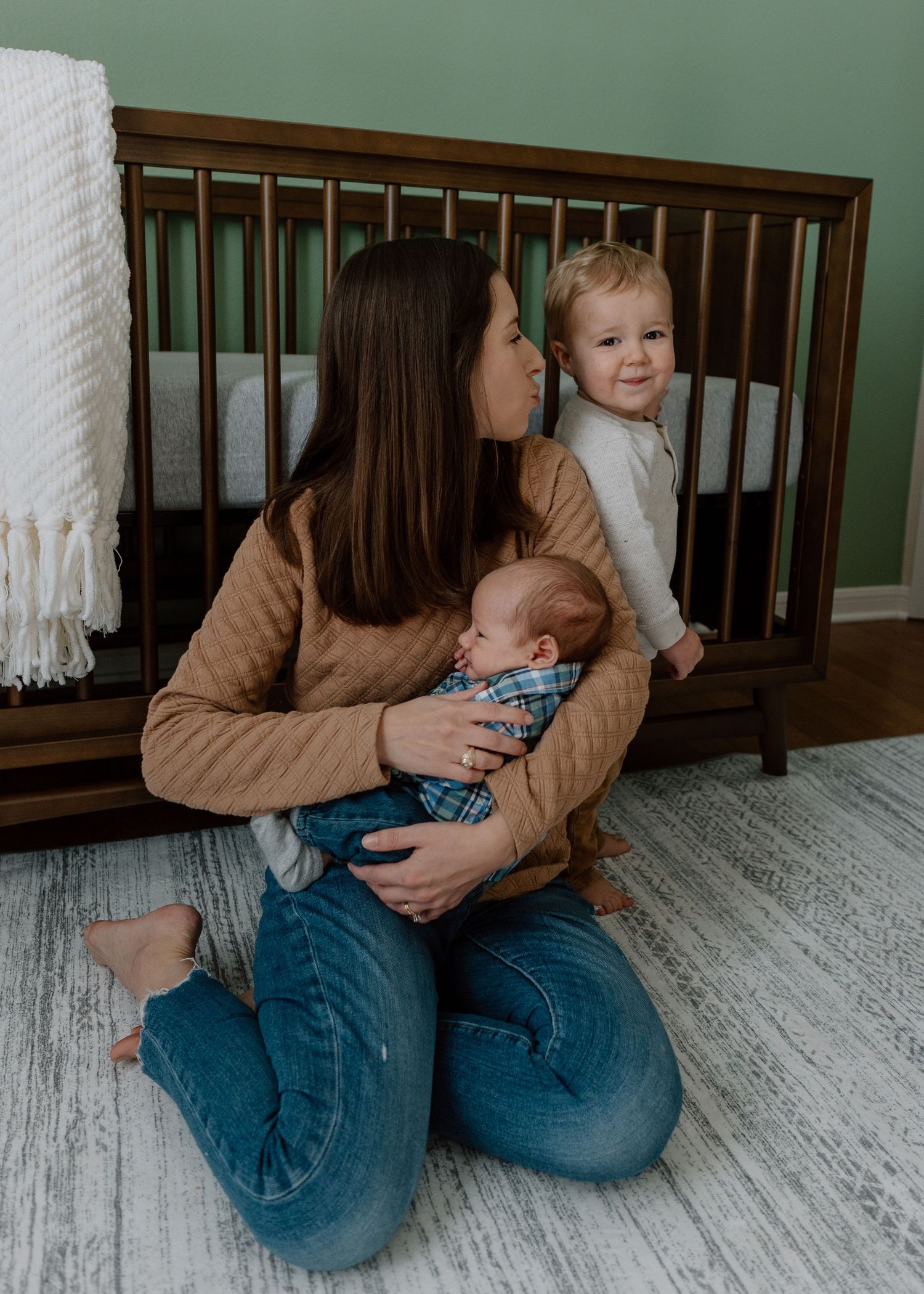 Mother in tan sweater and jeans kneels with two children next to wooden crib on light hardwood floor with green wall.