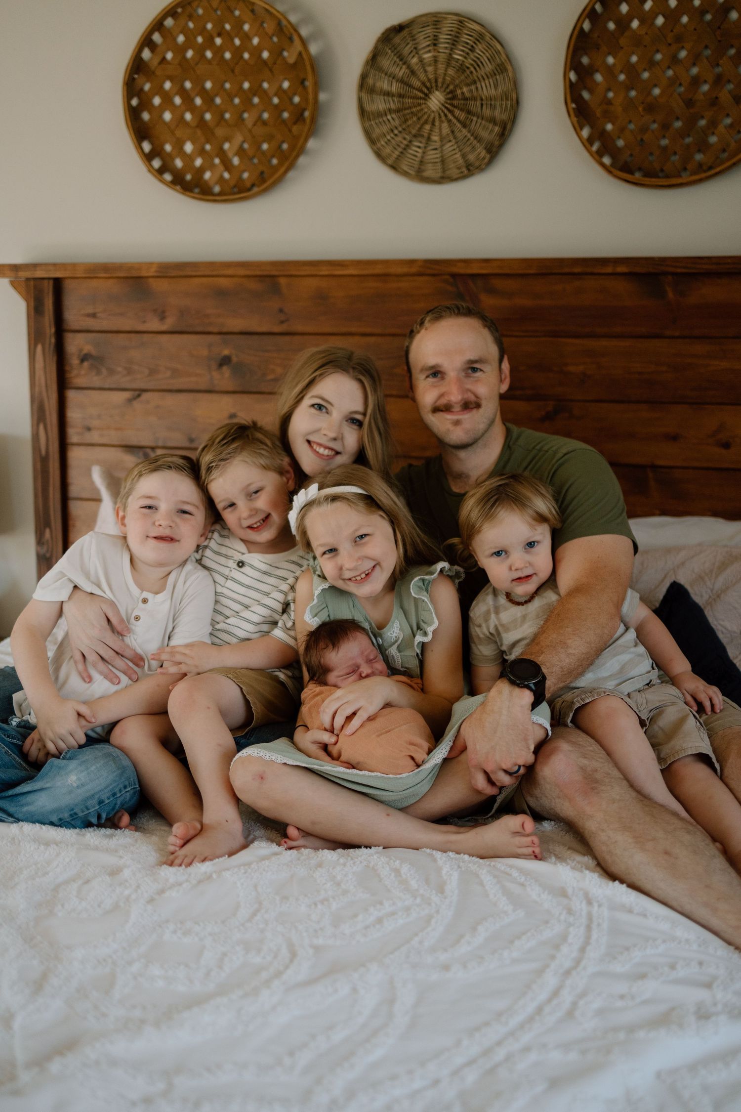 A family relaxes together on a bed with wooden headboard and decorative woven baskets displayed on the wall above.