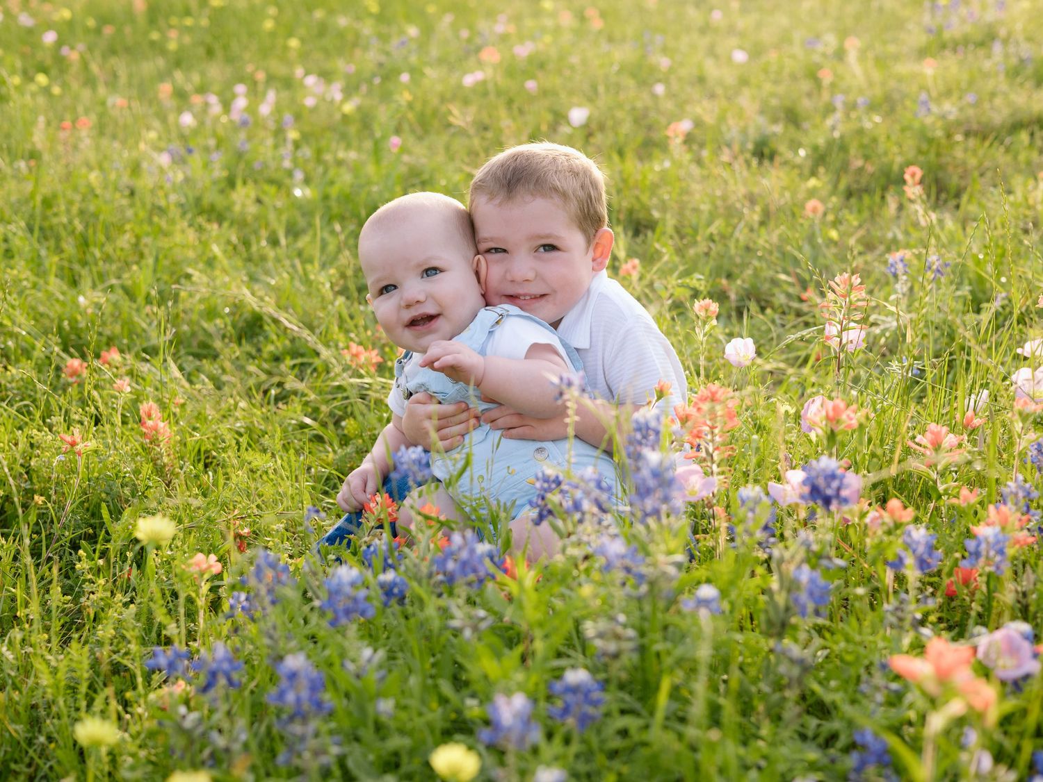 Two young children embrace while sitting in a wildflower meadow with purple and orange blooms in summer sunlight.