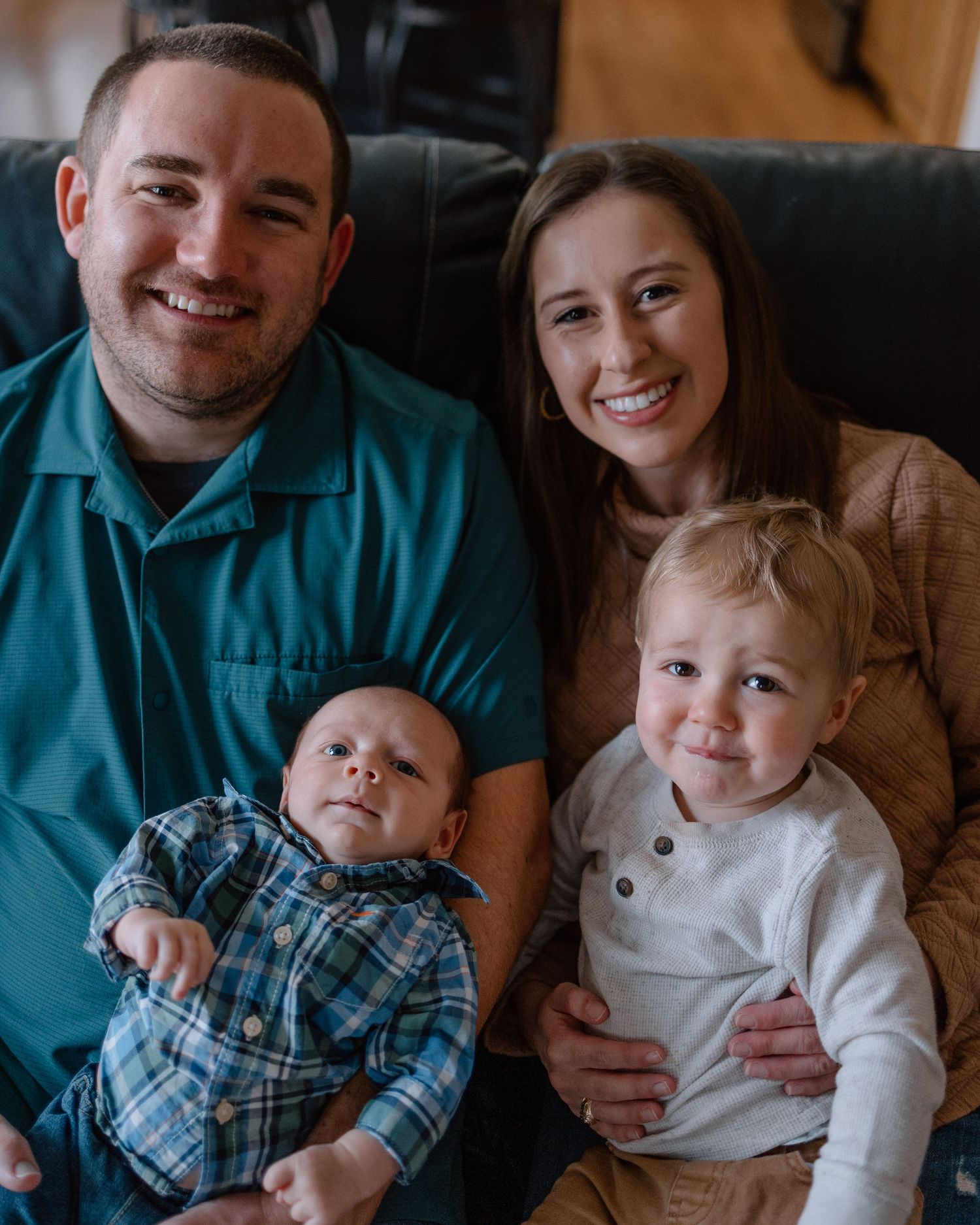 A family relaxes together on a couch, with two young children including a baby in plaid and a toddler in white.