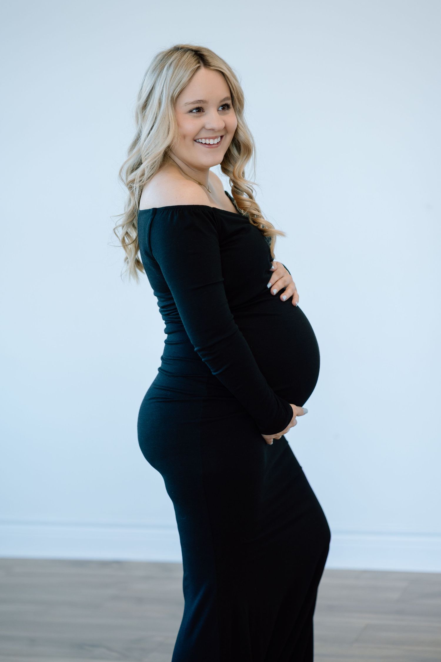 An expectant woman in an elegant black off-shoulder dress poses against a light background during a maternity photoshoot.