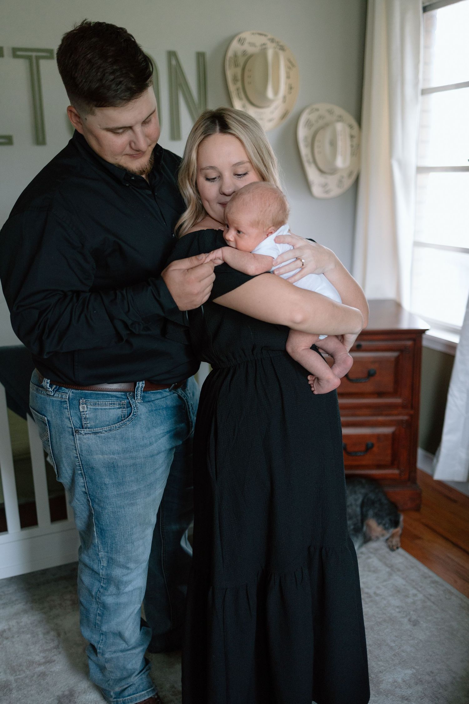 A couple stands in a cozy room with decorative plates on the wall while holding a newborn baby.