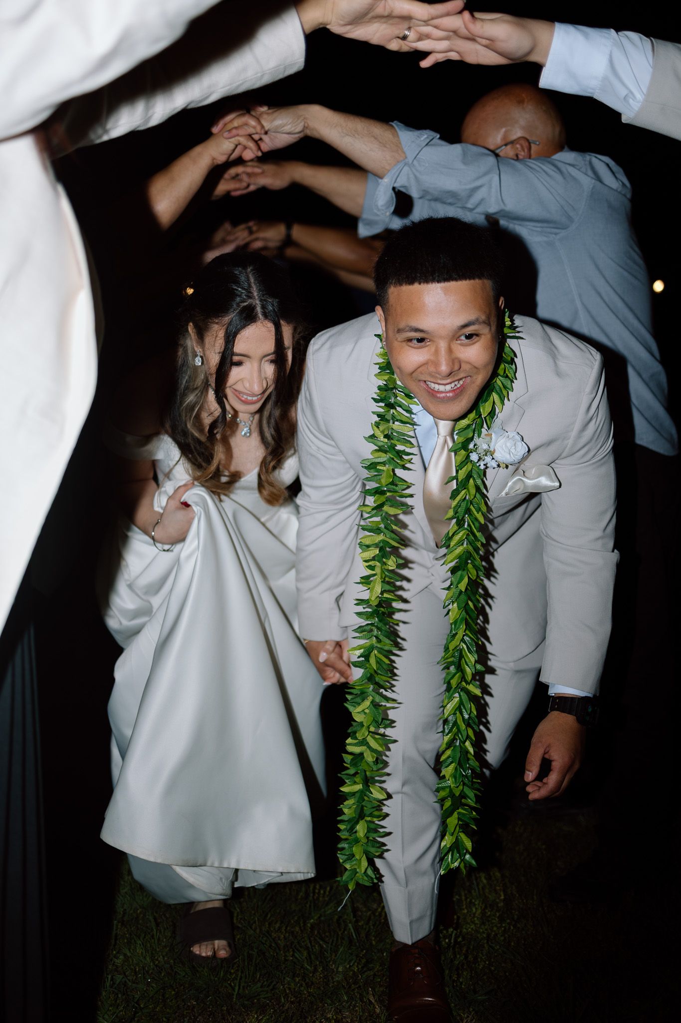 Wedding couple duck under guests' raised arms with groom wearing white suit and green Hawaiian lei.