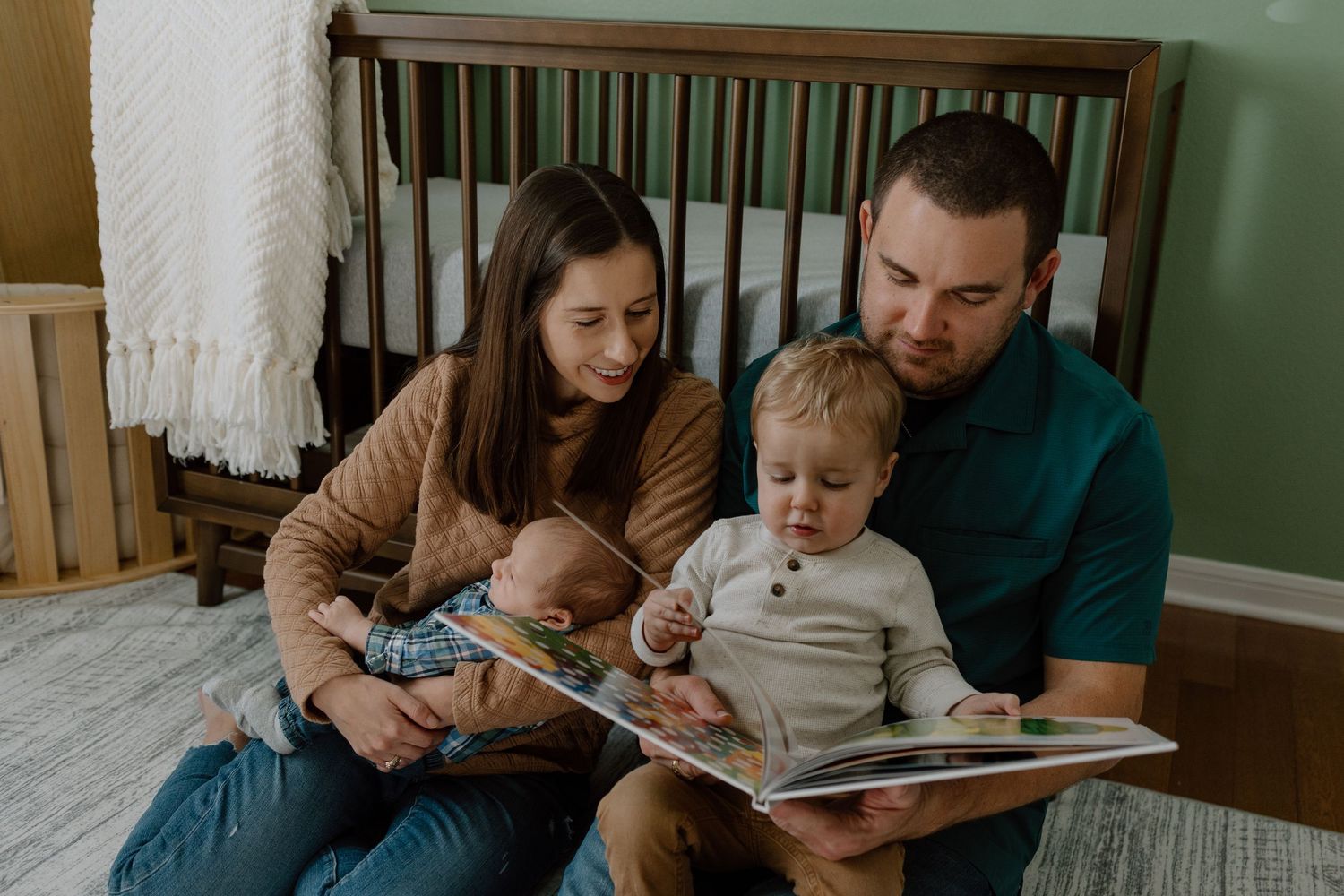 Family sitting on floor near wooden crib reading a picture book together in a cozy nursery room.