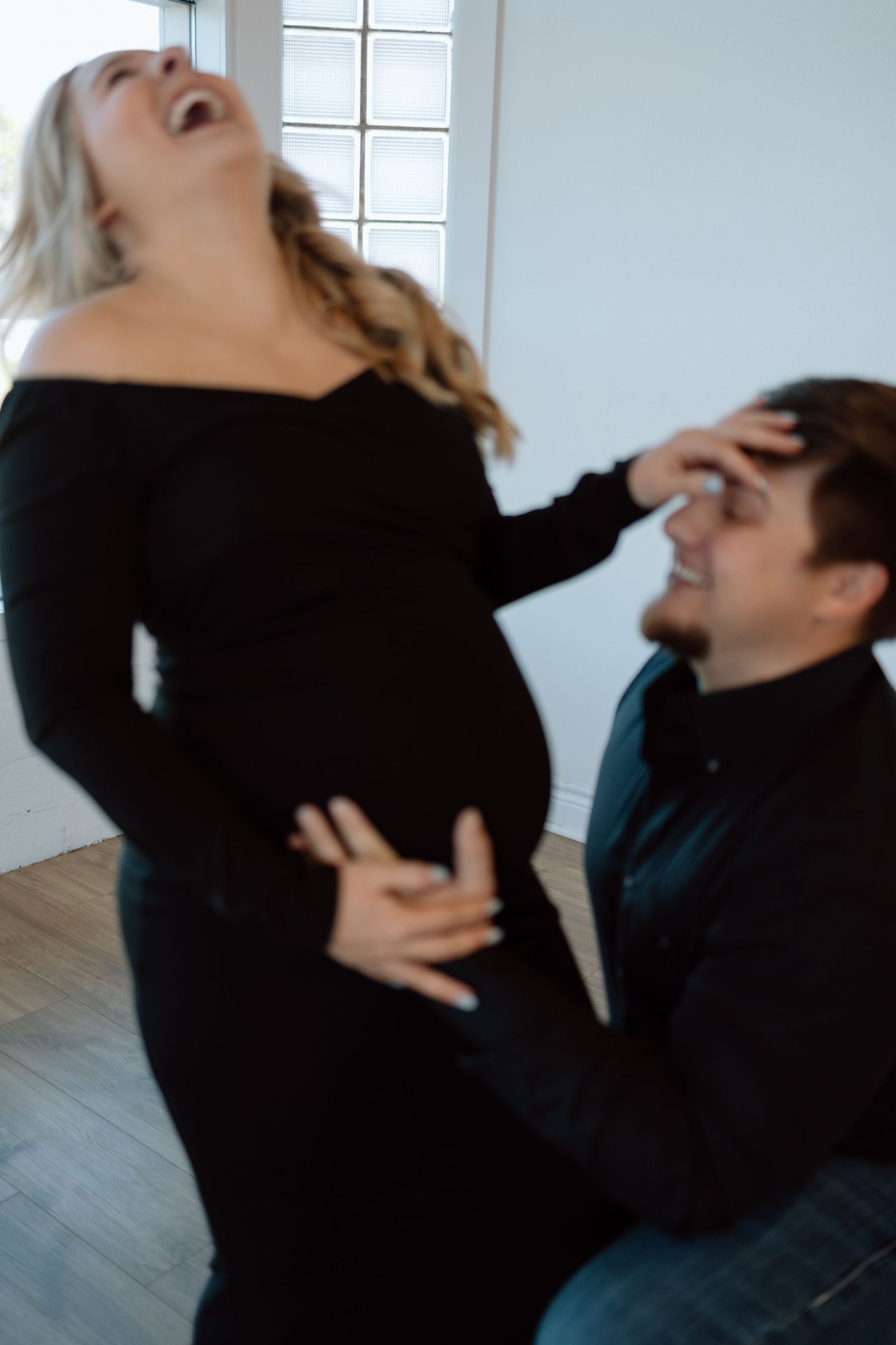 A couple shares a playful moment together as the woman in a black dress laughs while holding her pregnant belly.