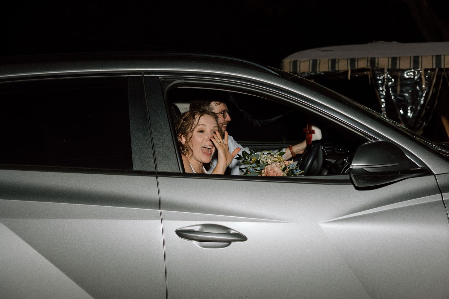 Newlyweds depart their wedding celebration in a silver car.