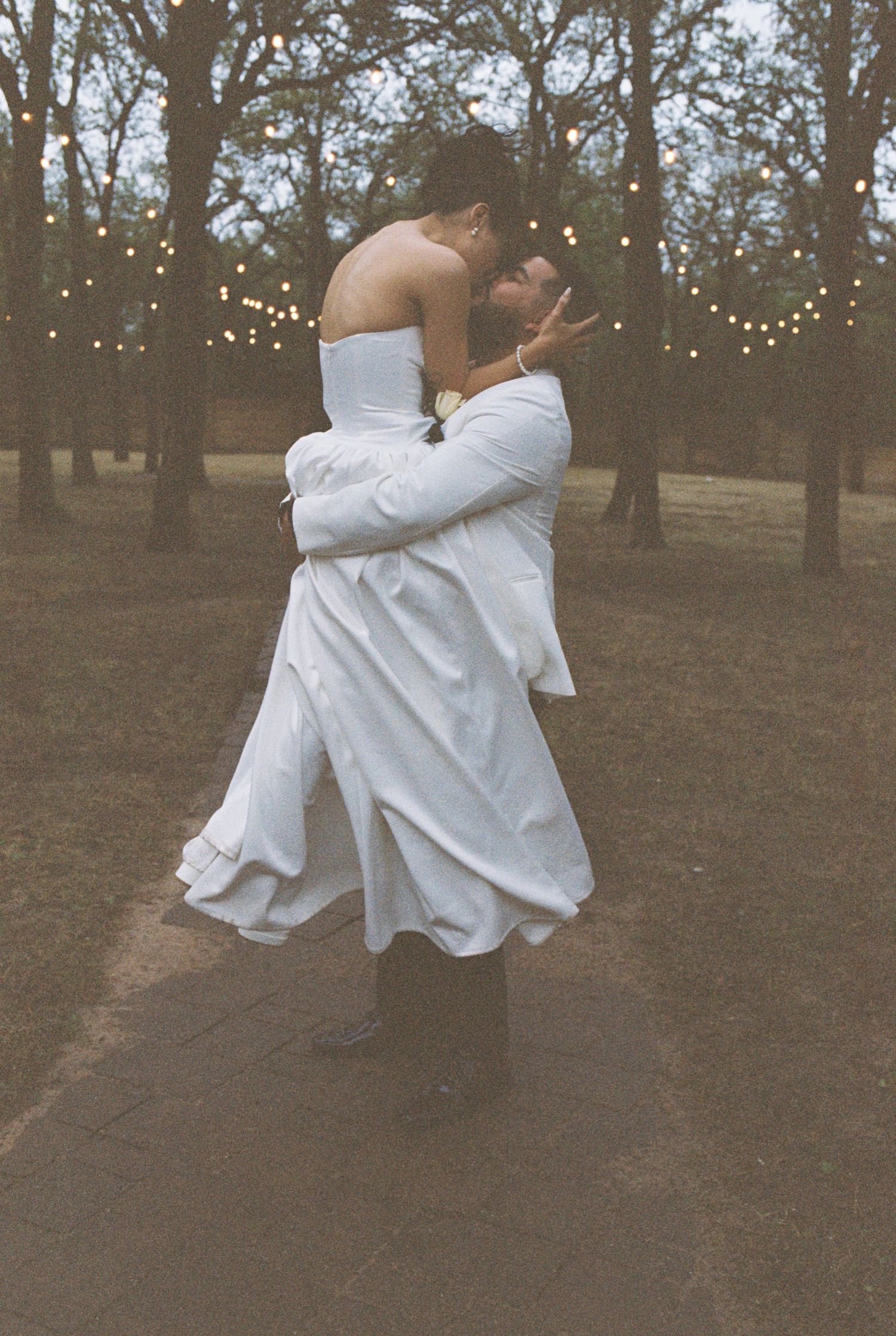 A romantic moment captured with fairy lights as a couple embraces in white attire in a rustic outdoor setting.