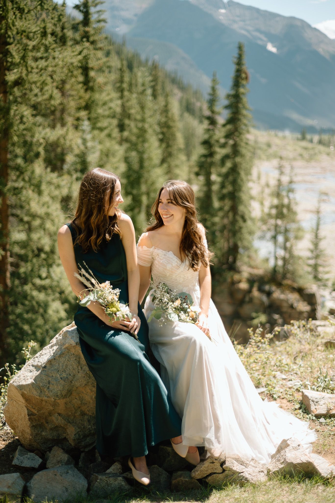 Two people in formal attire sit on rocks with mountain views and pine forests in the background.
