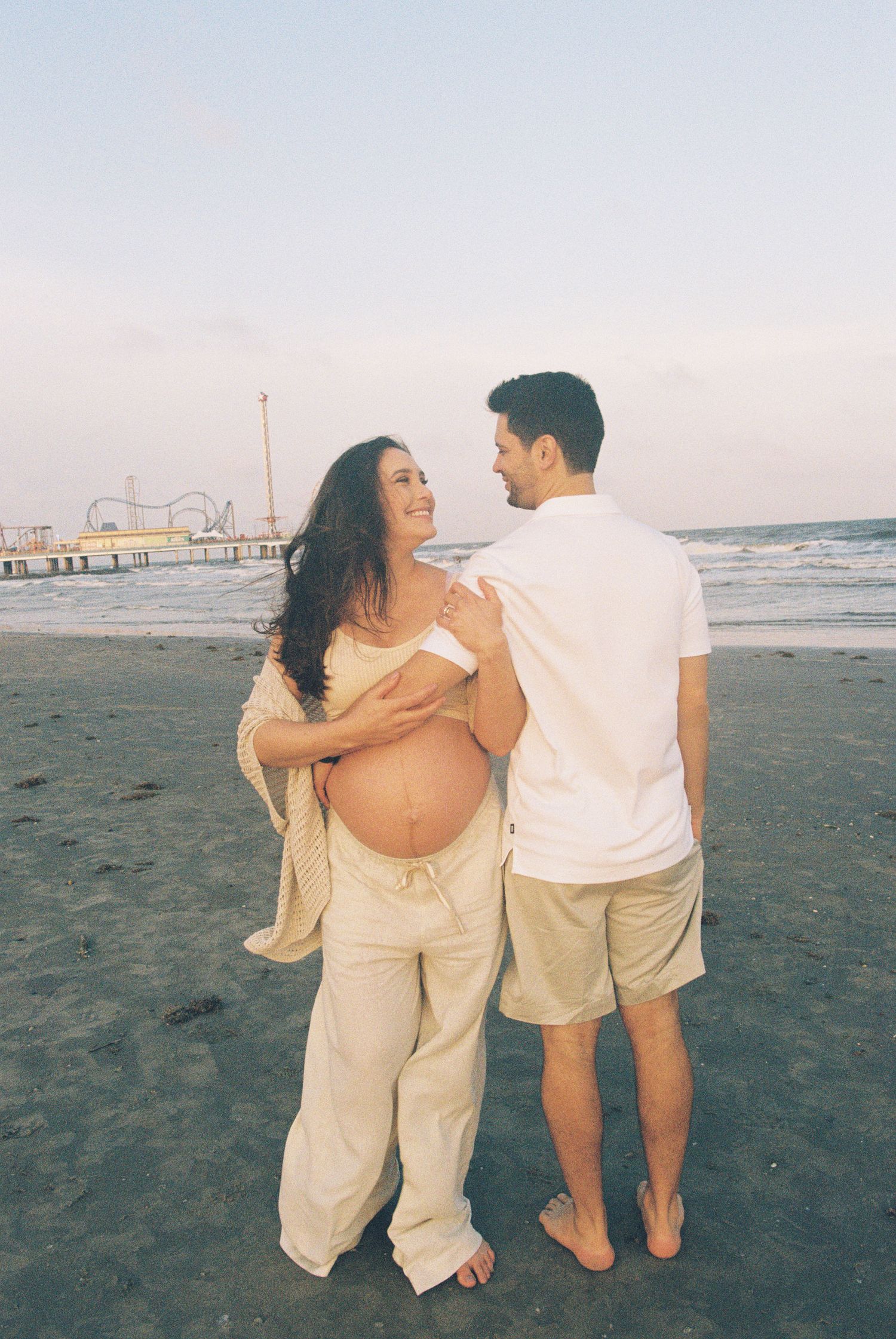 A couple shares a joyful moment on the beach with waves and pier in the background.