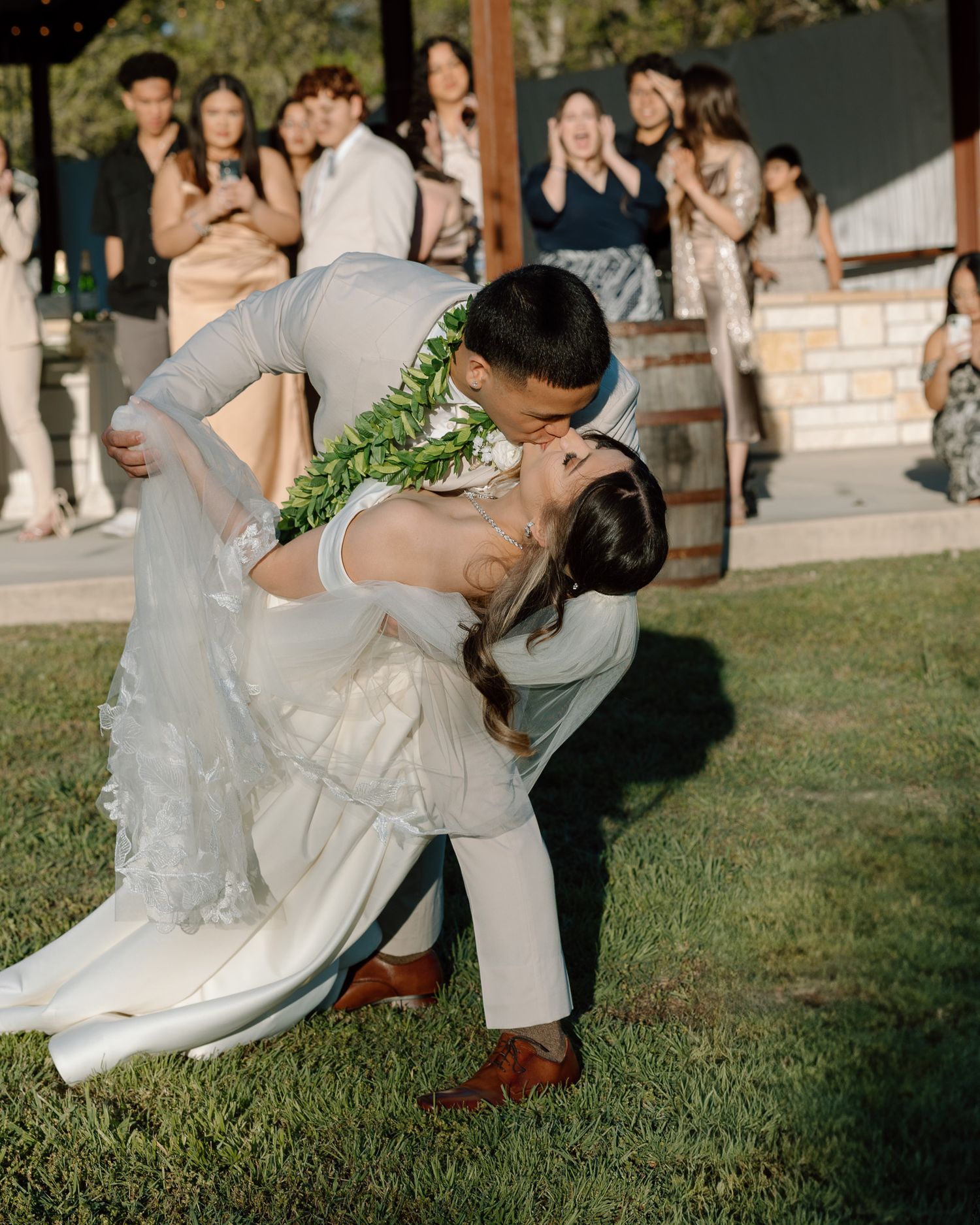 A romantic dip kiss on a grassy lawn with wedding guests in the background during an outdoor ceremony.