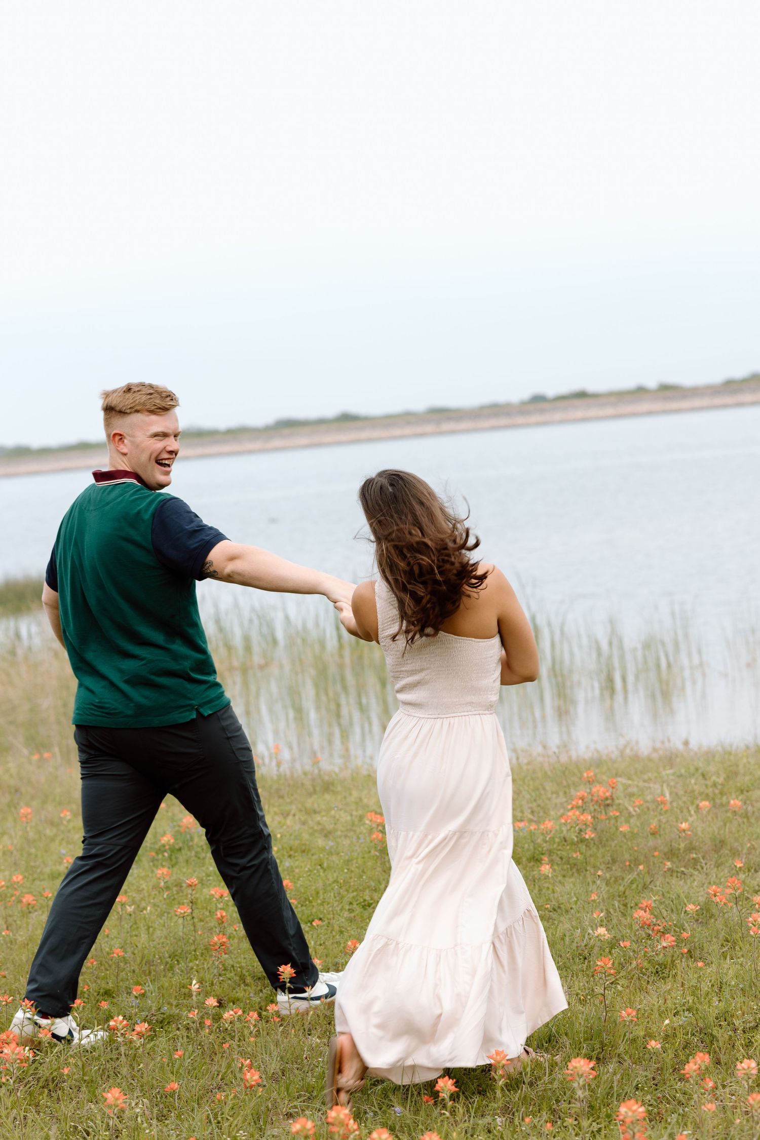 A couple runs playfully through a field of wildflowers near a lakeside.