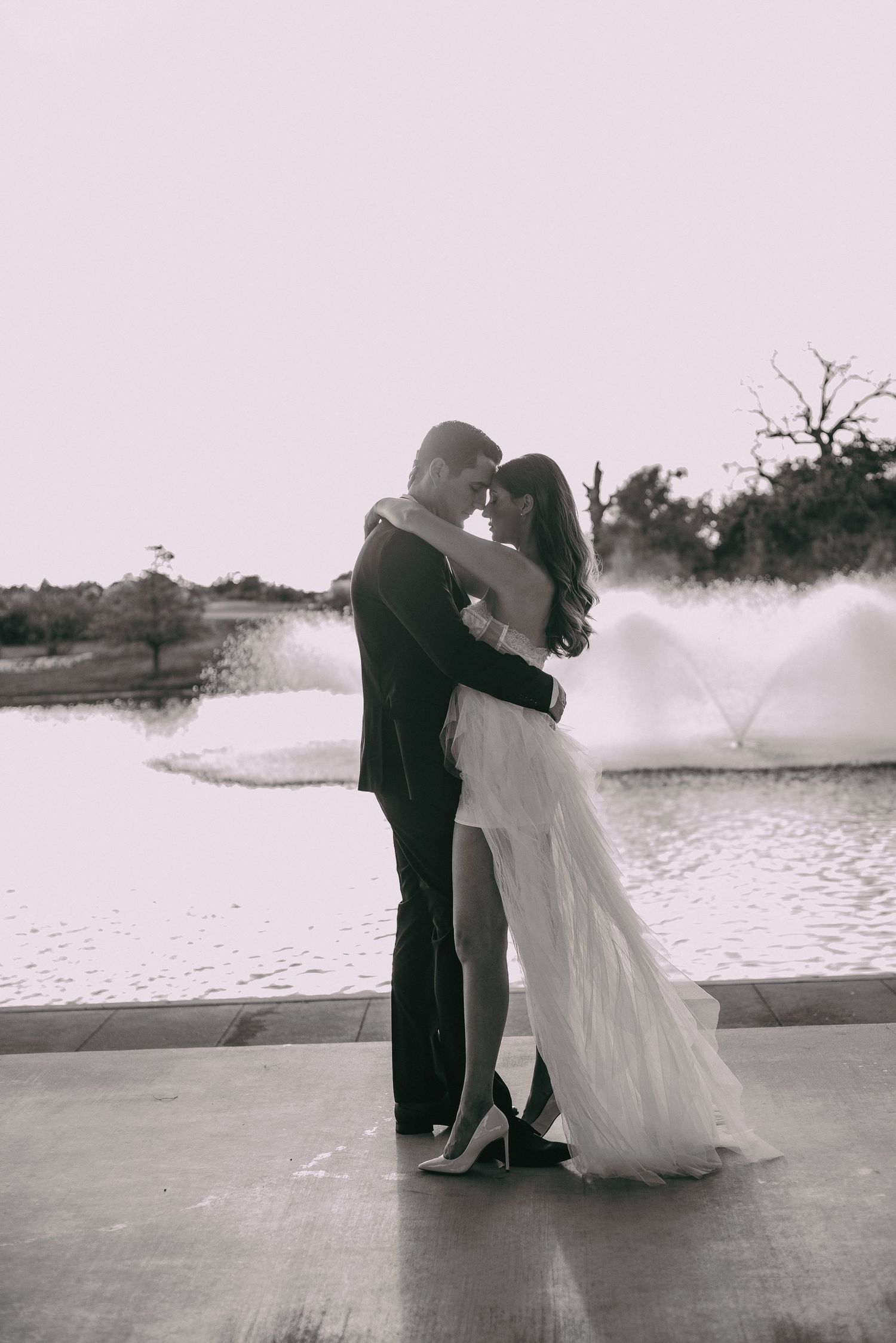 A romantic black and white portrait of a couple embracing on a deck near a fountain.