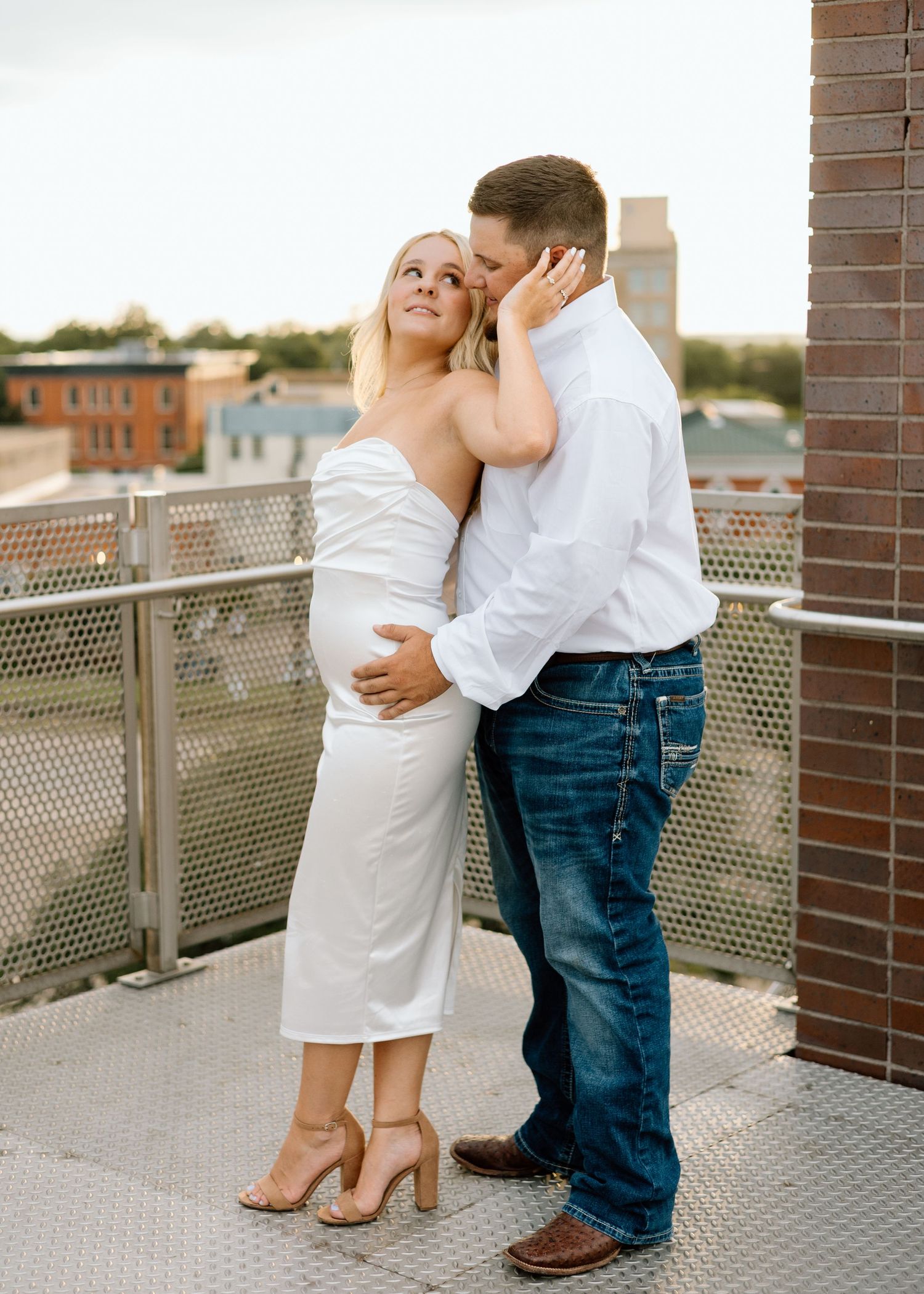 Pregnant woman in white dress embraced by man in white shirt and jeans on urban rooftop balcony.