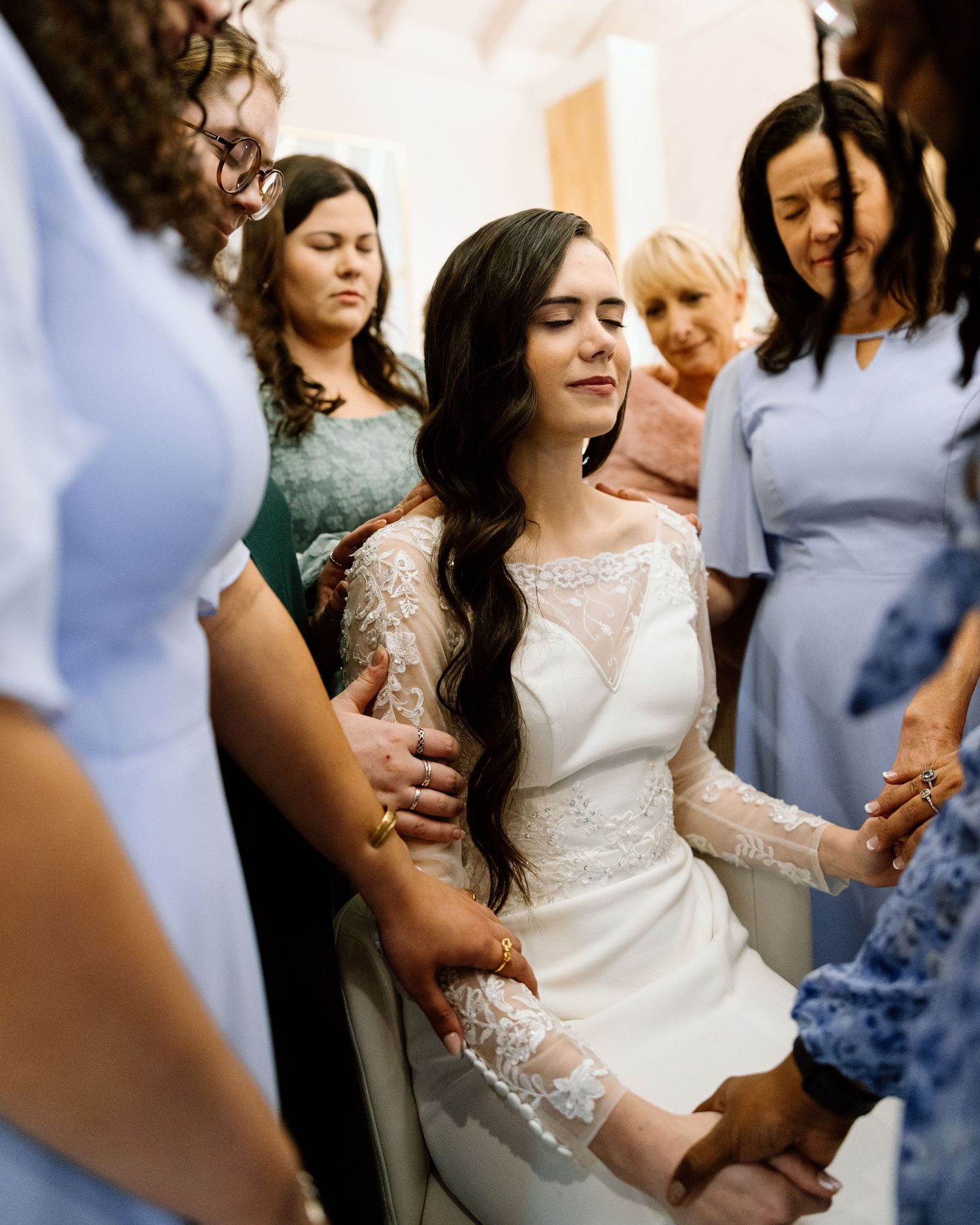 Emotional bride surrounded by loved ones in prayer circle before wedding ceremony.