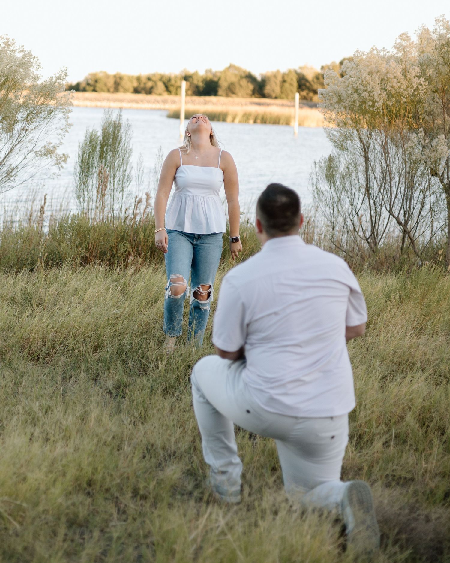 Person kneeling in grass proposing to a woman near a lake at golden hour, both wearing white.