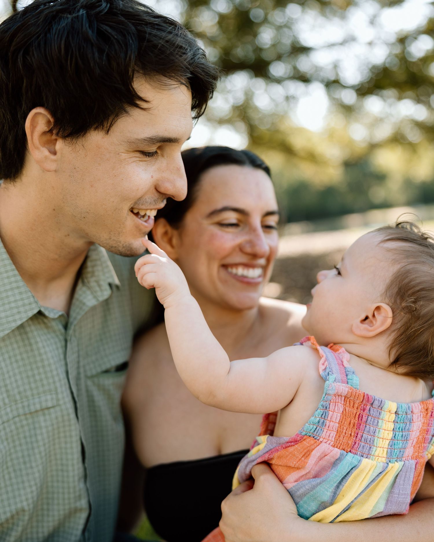 Smiling parents enjoy a sunny outdoor moment as their baby reaches up to touch dad's face.